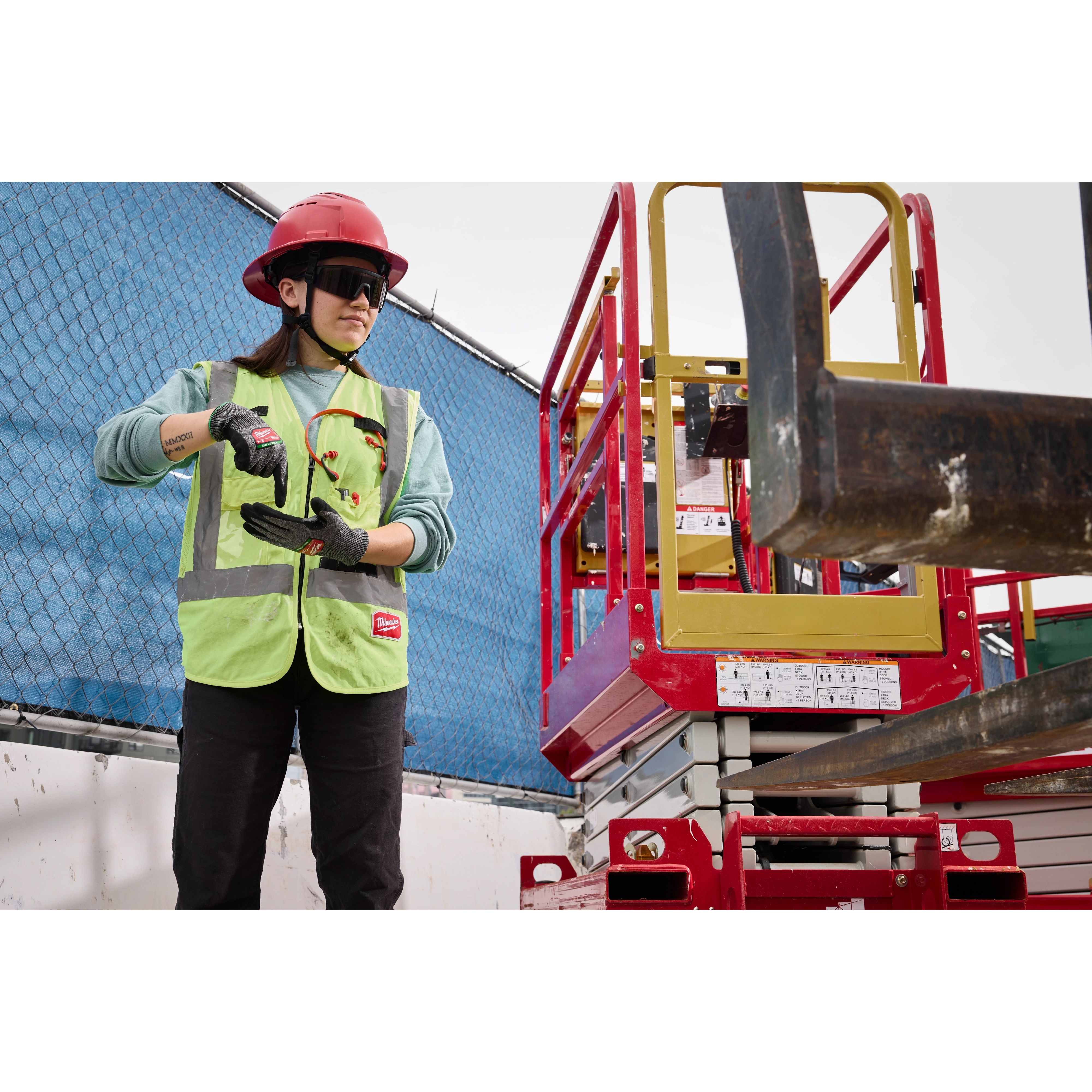 A construction worker wearing a red hard hat and neon green safety vest inspects a power tool on a site with a blue fence and red equipment in the background. The worker is wearing Wrap Around Safety Glasses - Tinted Anti-Scratch Lenses.