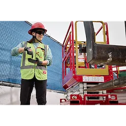 A construction worker wearing a red hard hat and neon green safety vest inspects a power tool on a site with a blue fence and red equipment in the background. The worker is wearing Wrap Around Safety Glasses - Tinted Anti-Scratch Lenses.