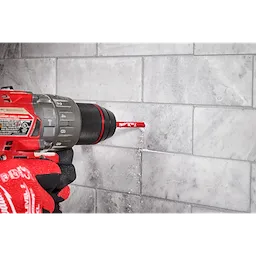 A Milwaukee® 1/4" DIAMOND MAX™ Hole Saw Bit is being used with a drill to cut into a tiled surface. A gloved hand is holding the drill steady as the drill bit creates a hole in the tiles. Dust and debris are visible around the drilling area.
