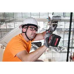 A construction worker in an orange shirt and white hard hat uses a cordless drill on a metal railing indoors. They are wearing Full Frame Safety Glasses with Removable Side Shields – Clear Anti-Scratch Lenses and protective gloves. The worker appears focused on the task at hand.