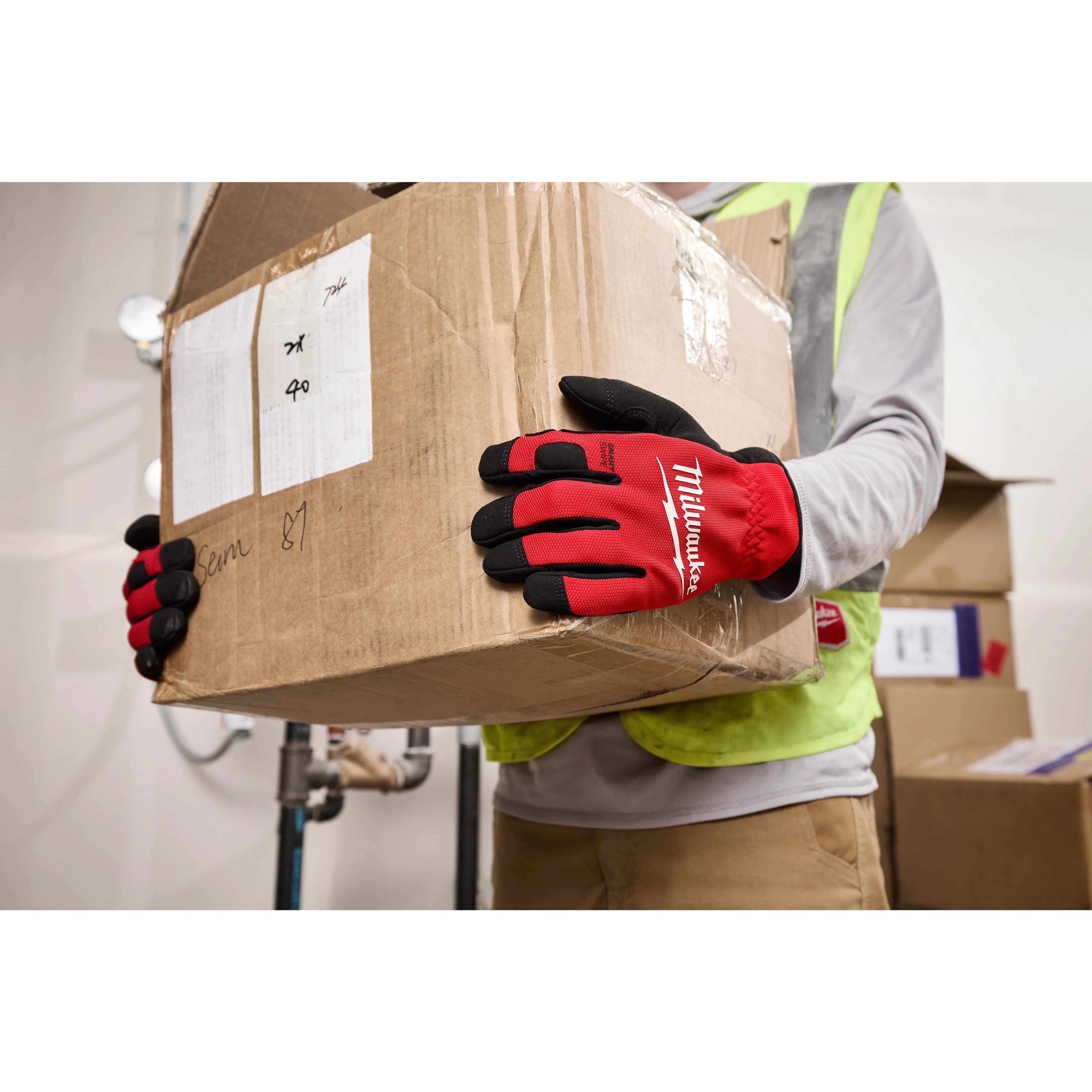 A person wearing Lightweight Work Gloves With Flexible Cuff is holding a large cardboard box. The gloves are red with black fingertips and have "Milwaukee" printed on the back. The individual is also wearing a yellow safety vest.