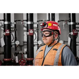 A worker wearing Non-Vented Goggles - Clear Dual Coat Lens, a red hard hat with a light, and an orange safety vest in an industrial setting.
