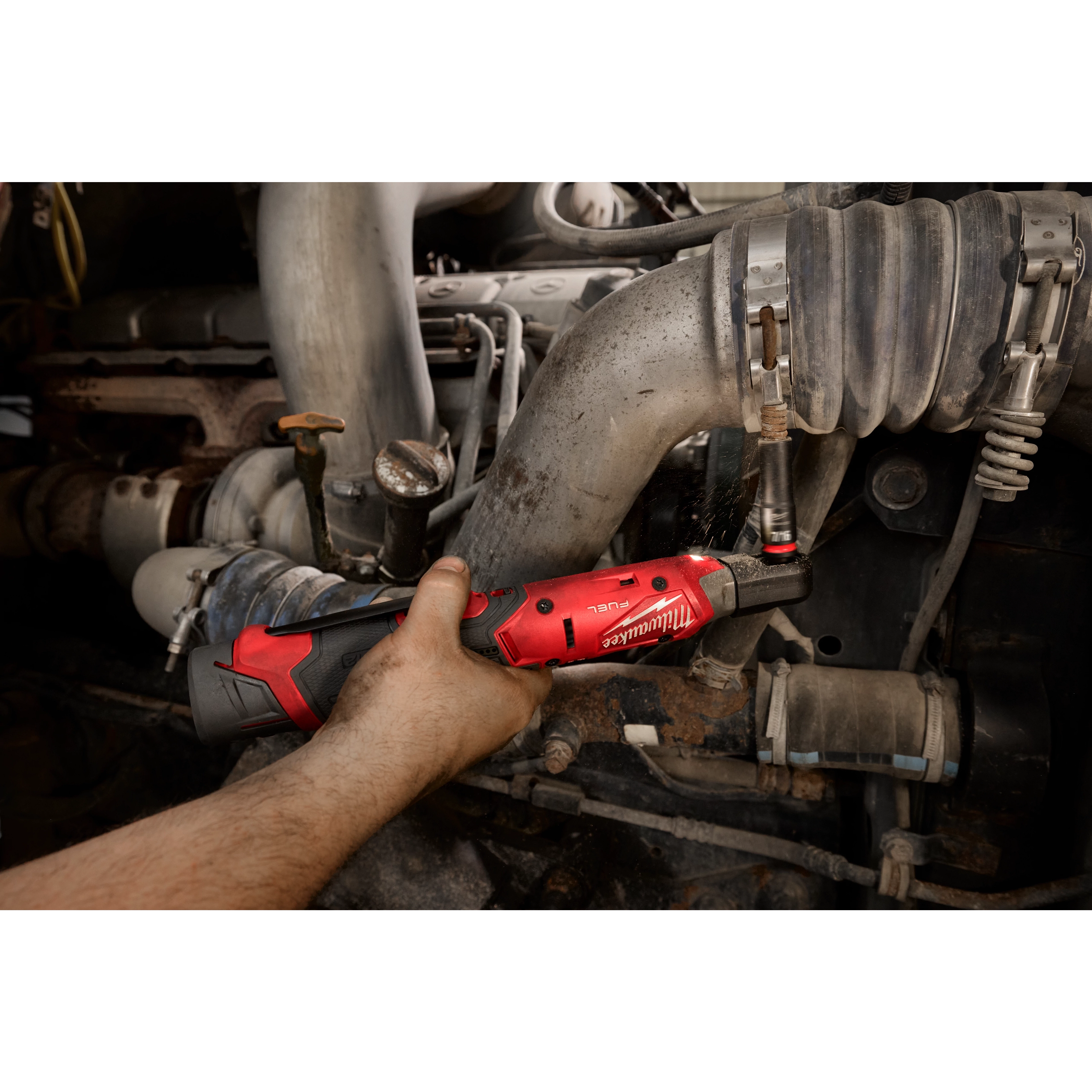A worker uses the M12 FUEL™ 3/8" Ratchet Kit to tighten a nut in a mechanic shop. The tool has a red body with black accents and an ergonomic handle, designed for automotive maintenance and repairs in tight spaces. The background shows engine parts and hoses.