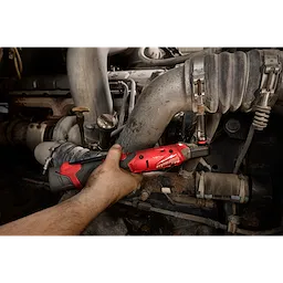 A worker uses the M12 FUEL™ 3/8" Ratchet Kit to tighten a nut in a mechanic shop. The tool has a red body with black accents and an ergonomic handle, designed for automotive maintenance and repairs in tight spaces. The background shows engine parts and hoses.