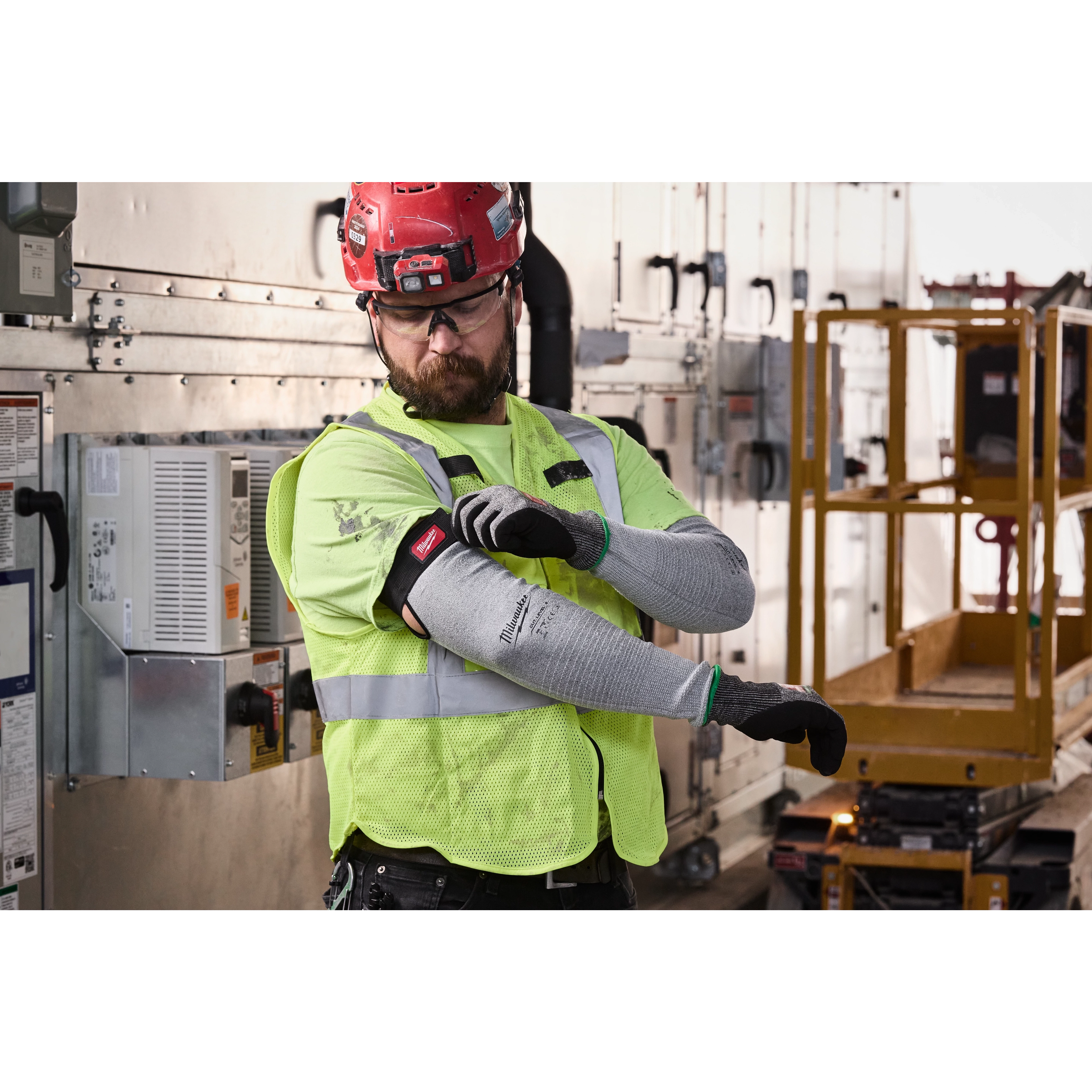 Worker wearing high-visibility vest adjusts Cut Level 6 Protective Sleeves in an industrial setting with control panels and scaffold.