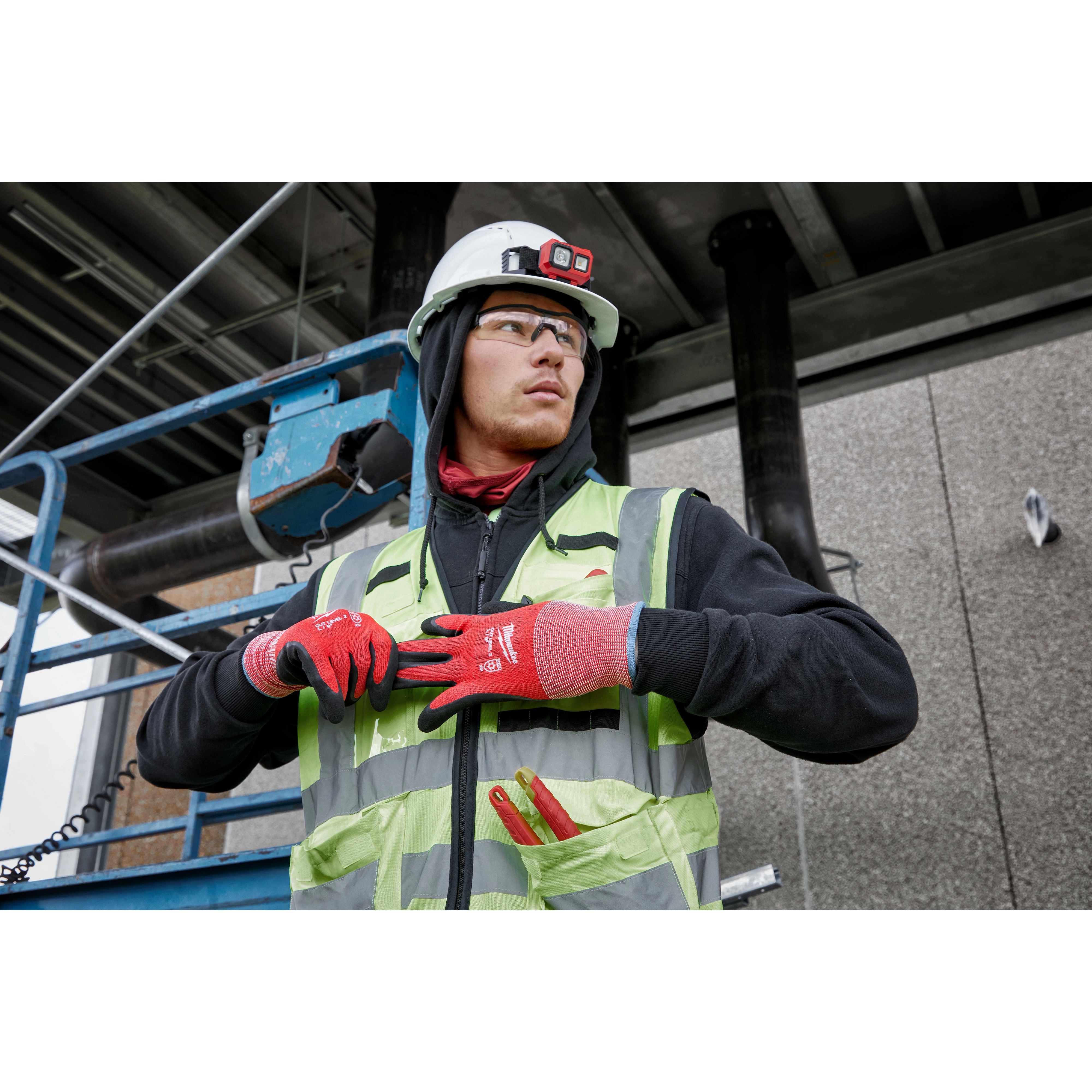 A construction worker wearing a white hard hat and high-visibility vest is adjusting a strap. The worker's red gloves are prominently visible, displaying "Cut Level 2 Winter Dipped Gloves - L." A utility knife is shown in the vest pocket. The worker is positioned in an industrial site.