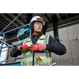 A construction worker wearing a white hard hat and high-visibility vest is adjusting a strap. The worker's red gloves are prominently visible, displaying "Cut Level 2 Winter Dipped Gloves - L." A utility knife is shown in the vest pocket. The worker is positioned in an industrial site.