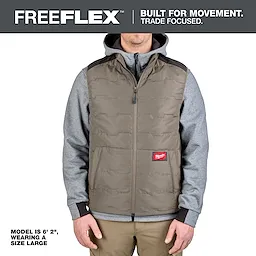 Man wearing a brown FREEFLEX™ Lightweight Insulated Vest over a gray hoodie. The vest has a zip front and a red logo patch on the lower left side. Text indicates the model is 6' 2", wearing a size large. The slogan above reads "Built for movement. Trade focused."