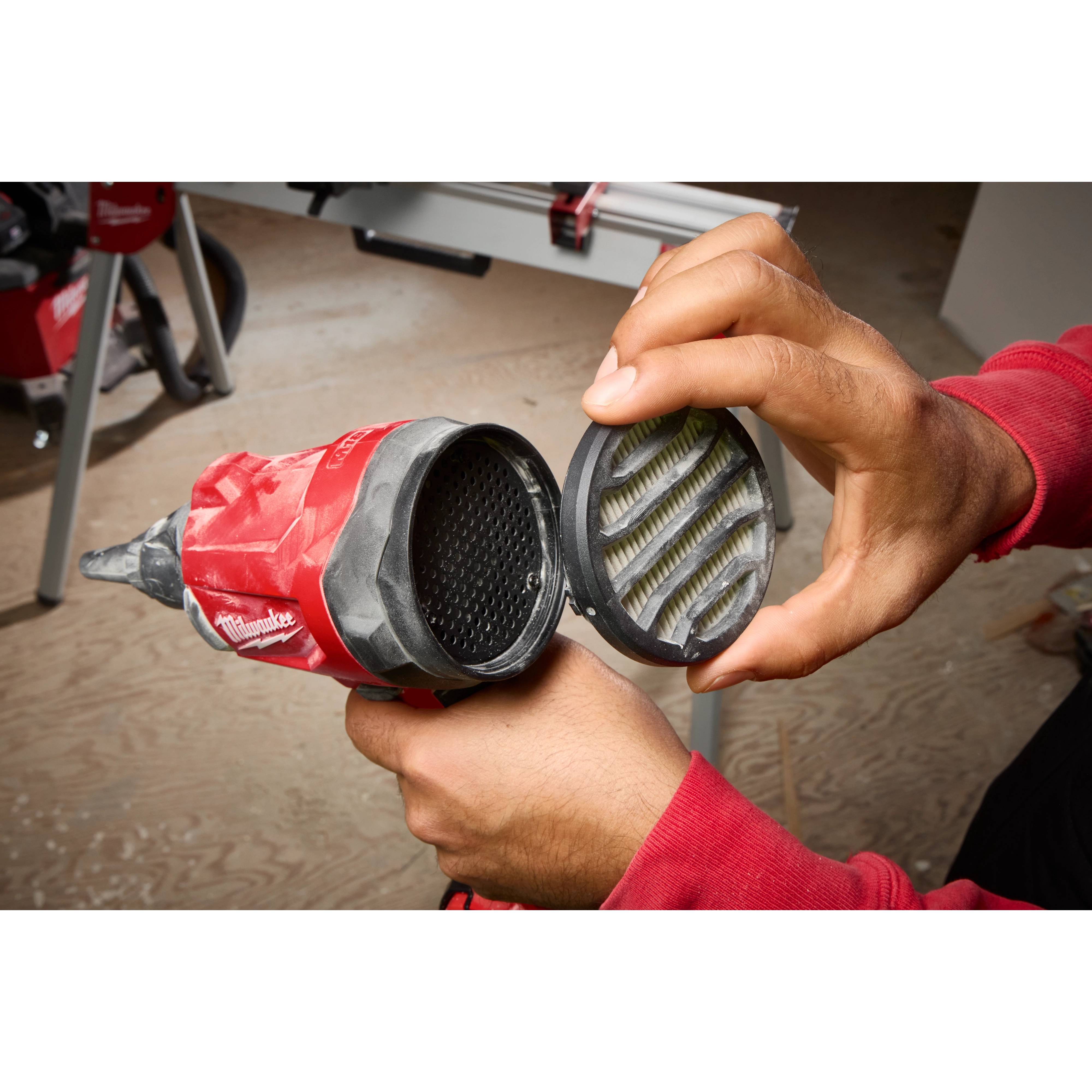 A person in a red shirt holds the M18™ Brushless Precision Blower with one hand while removing its filter cover with the other. The tool is red and black with a visible interior filter.