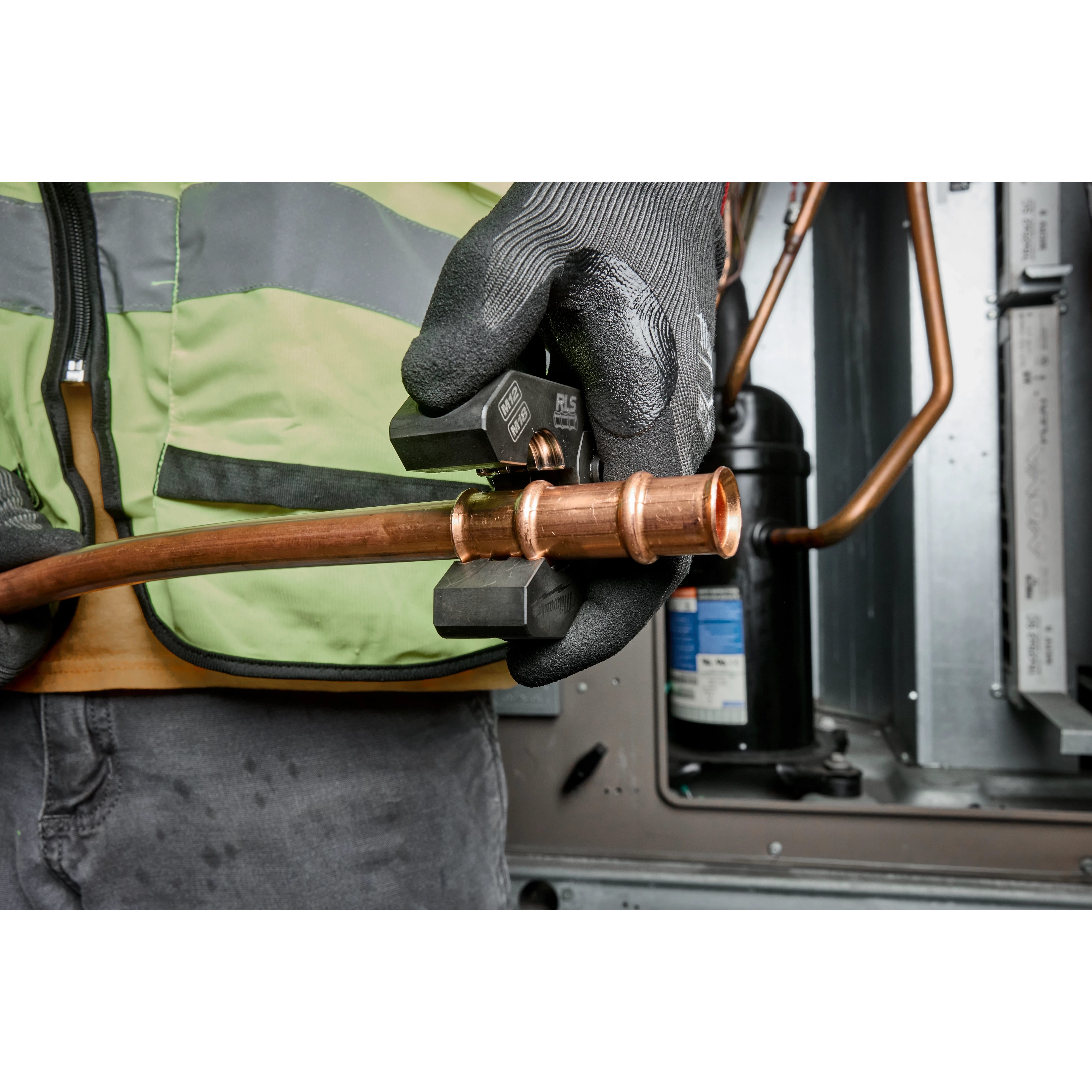 A worker wearing gloves uses the 1/4" - 7/8" RLS® Pivoting Press Ring Kit for M12™ FORCE LOGIC™ Press Tool on copper tubing in an HVAC setting. The worker is wearing a green safety vest while operating the tool.