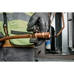A worker wearing gloves uses the 1/4" - 7/8" RLS® Pivoting Press Ring Kit for M12™ FORCE LOGIC™ Press Tool on copper tubing in an HVAC setting. The worker is wearing a green safety vest while operating the tool.