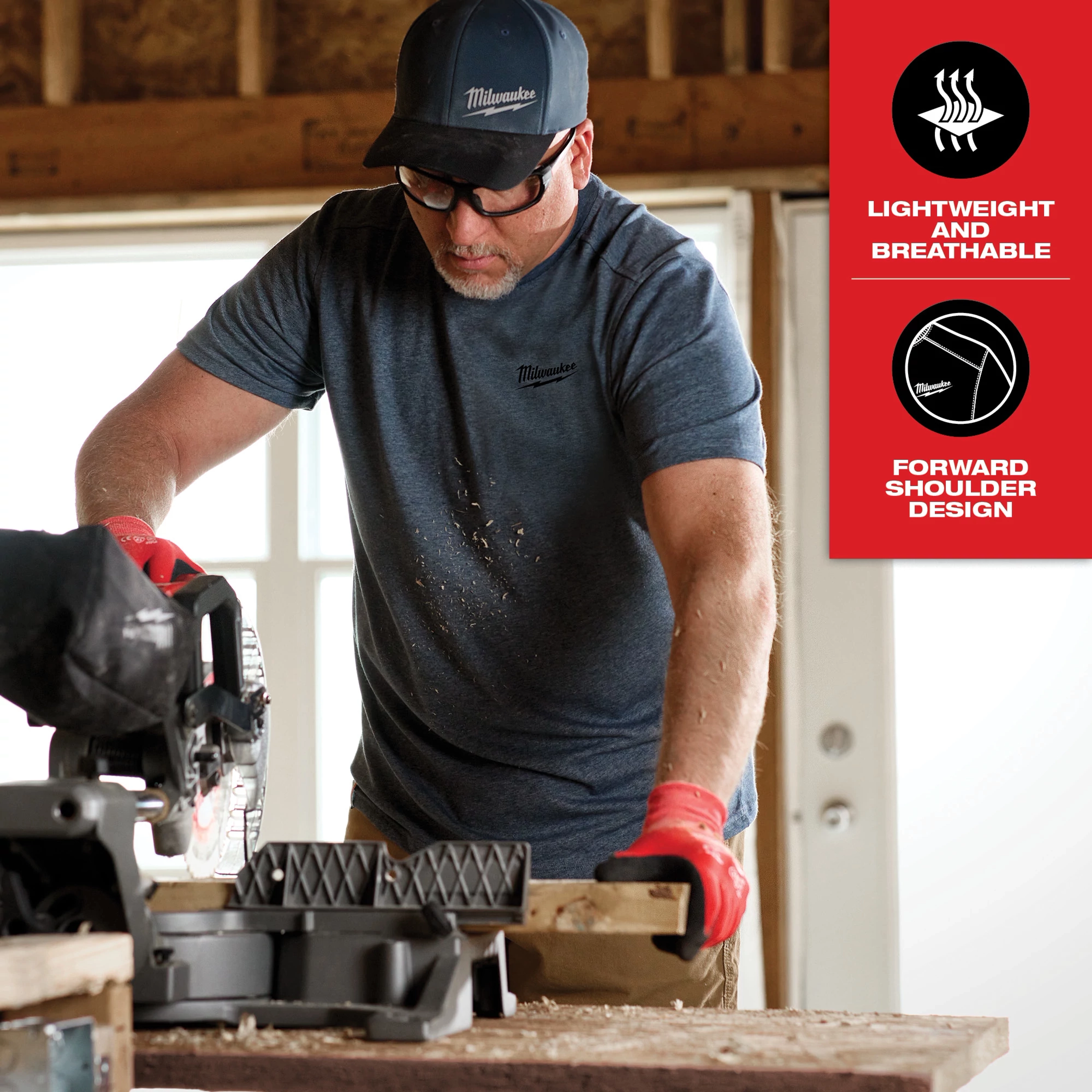 A man wearing a blue Hybrid Work Tee - Short Sleeve operates a saw in a workshop. The tee is described as lightweight and breathable with a forward shoulder design.