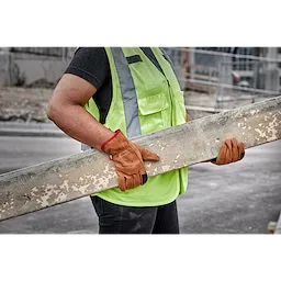 Person wearing Goatskin Leather Gloves holding a wooden plank, dressed in a green safety vest and gray shirt at a construction site.