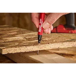 A hand holds the INKZALL™ 2IN1 Deep Reach Ultra Fine Point Black Jobsite Marker, marking a piece of wood with precision. The marker features a red body and a black fine point tip, ideal for detailed work on rough surfaces.