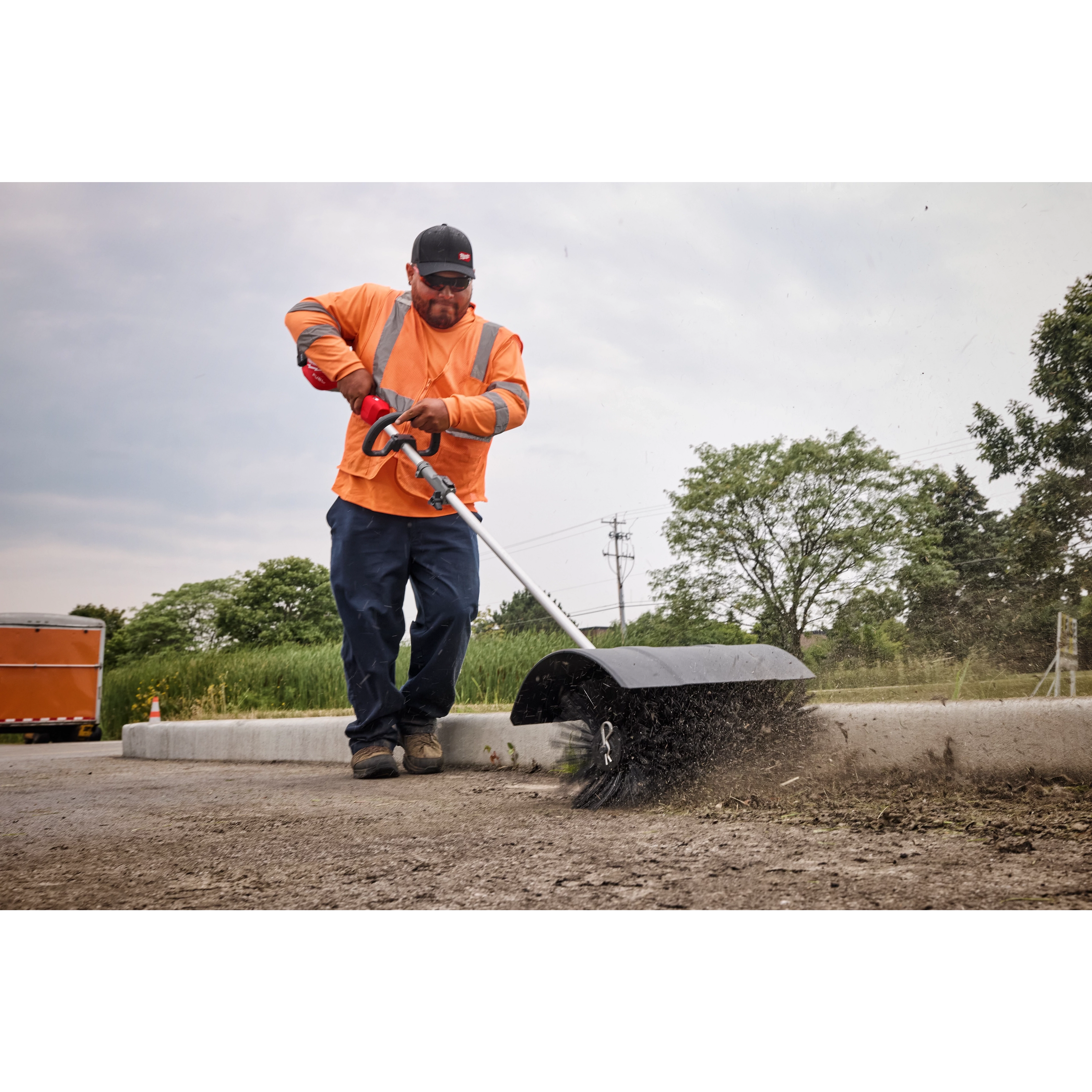 Worker in an orange safety vest using a street sweeper on a road, surrounded by greenery and traffic cones.