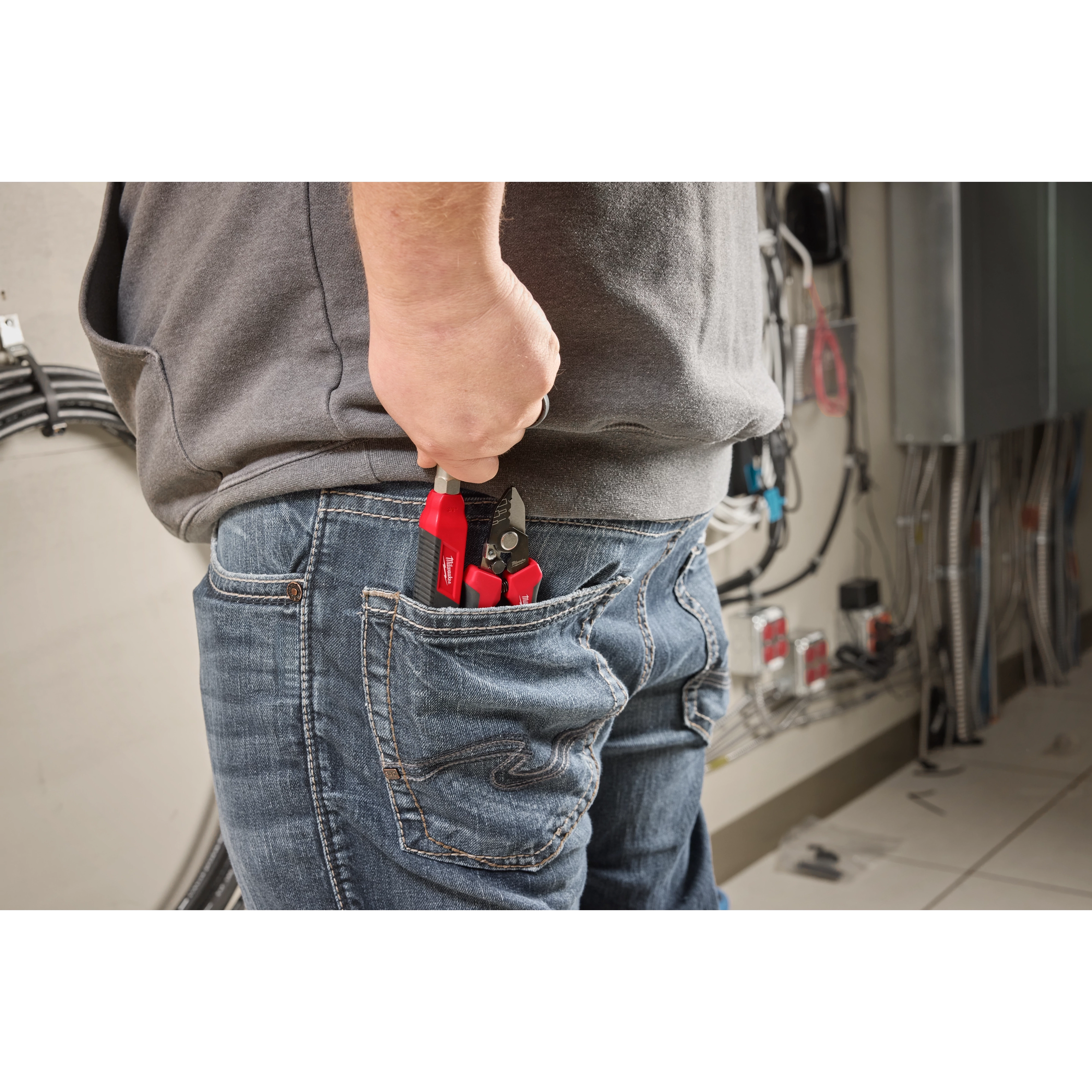 A person in a gray shirt and jeans holds a Can Wrench (3/8” & 7/16”) partially tucked in their back pocket while standing in an industrial setting with cables and electrical components visible in the background.