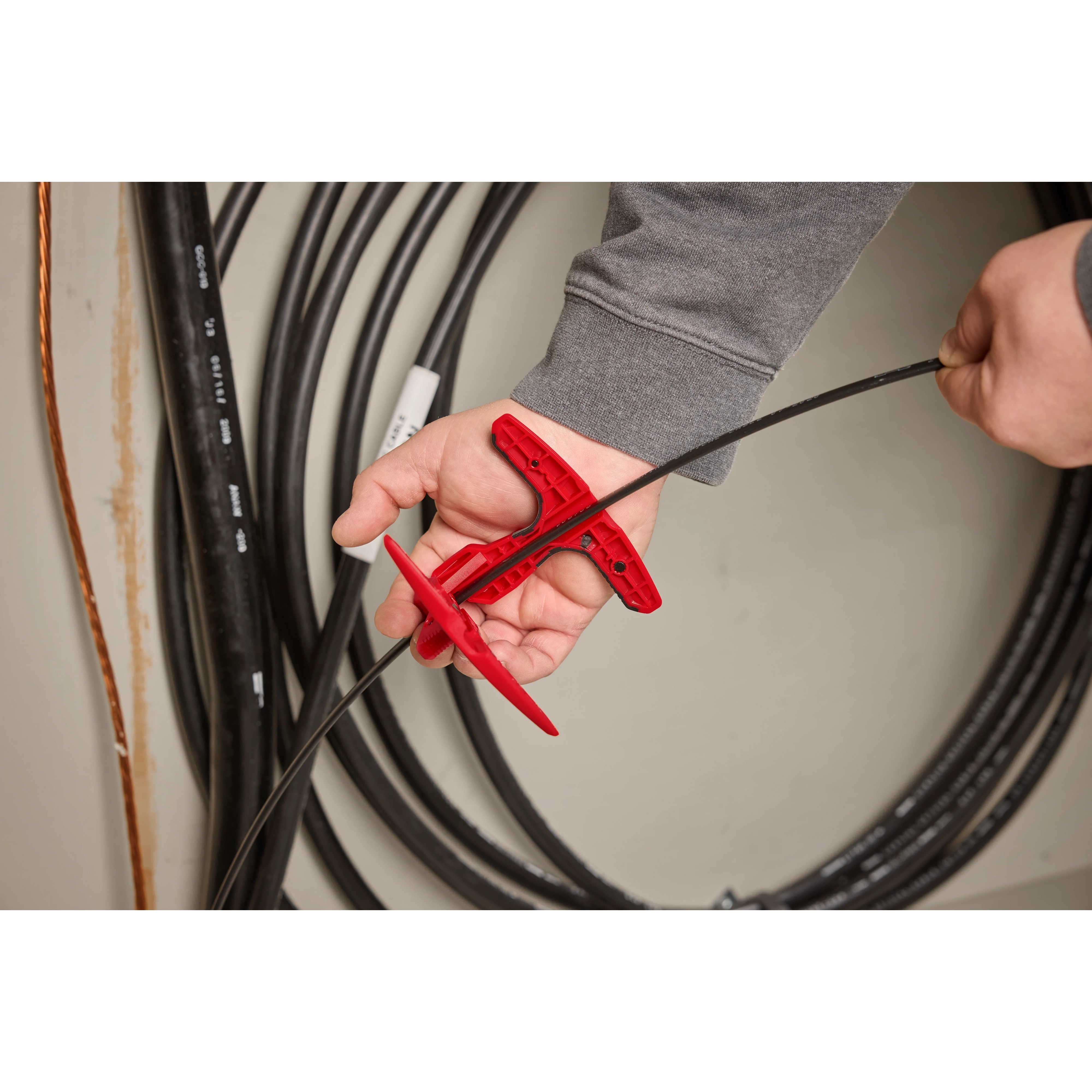 A person uses a red Fiber Optic Drop Cable Slitter to cut a black fiber optic cable. Coils of cables can be seen in the background.