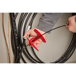 A person uses a red Fiber Optic Drop Cable Slitter to cut a black fiber optic cable. Coils of cables can be seen in the background.