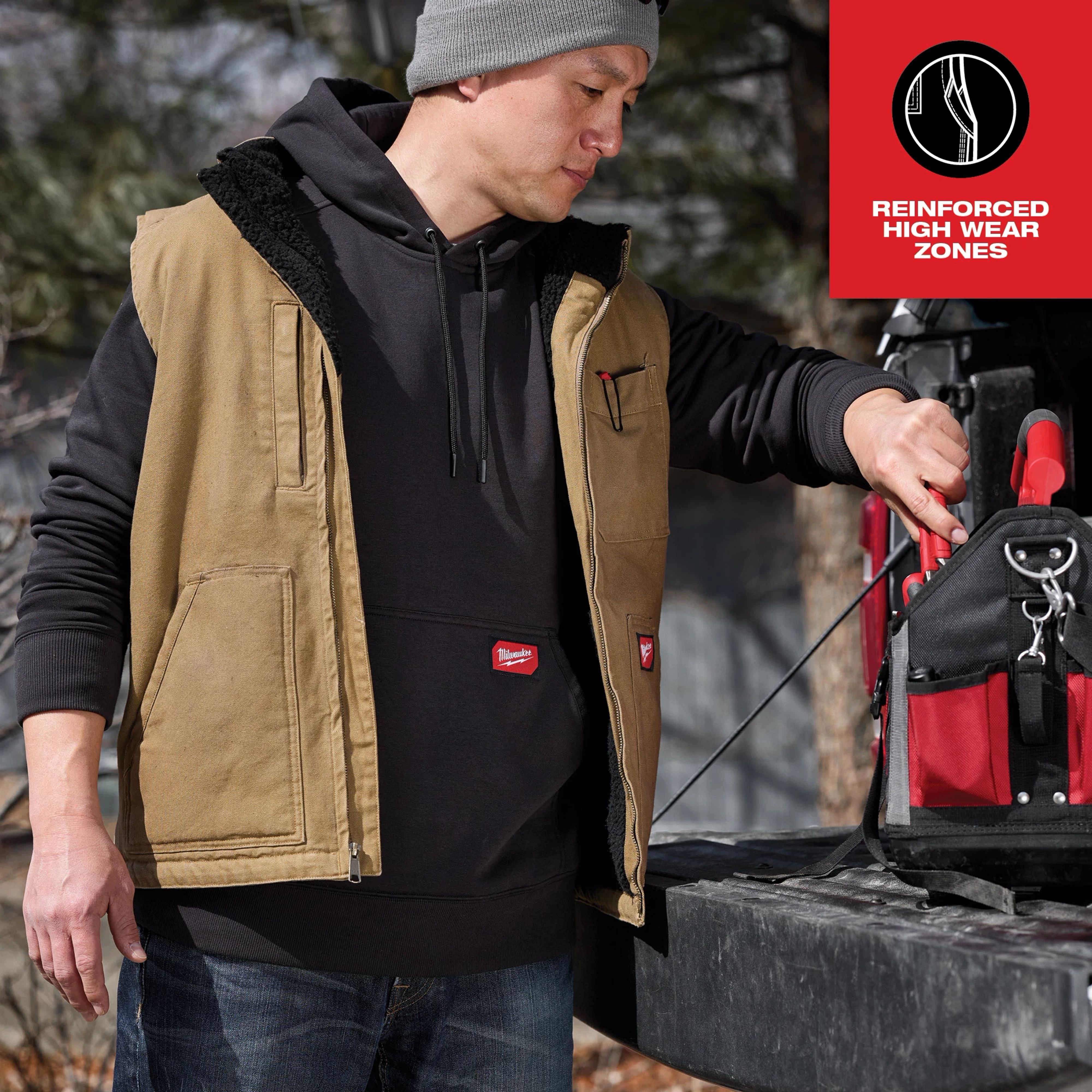 A person wearing a Heavy Duty Sherpa-Lined Vest Feature Shot in khaki color, standing beside a red and black tool bag on a truck.
