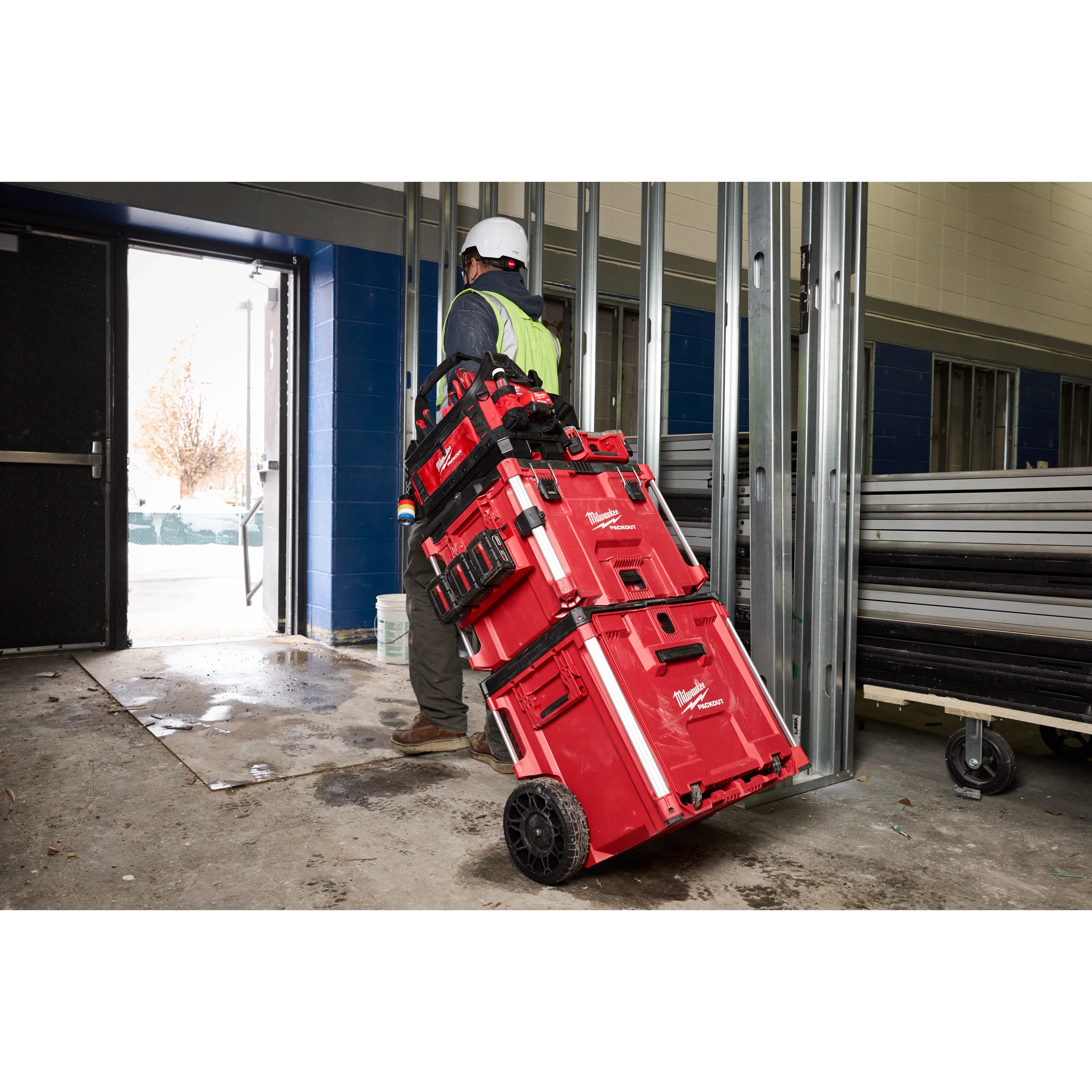 Worker with hard hat and safety vest moving PACKOUT XL Tool Box stack on wheels through a construction site doorway.