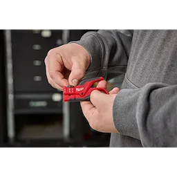 A person uses a red Buffer Tube Ringer & Slitter tool to strip cables. They are holding the tool in both hands, with a gray jacket visible.