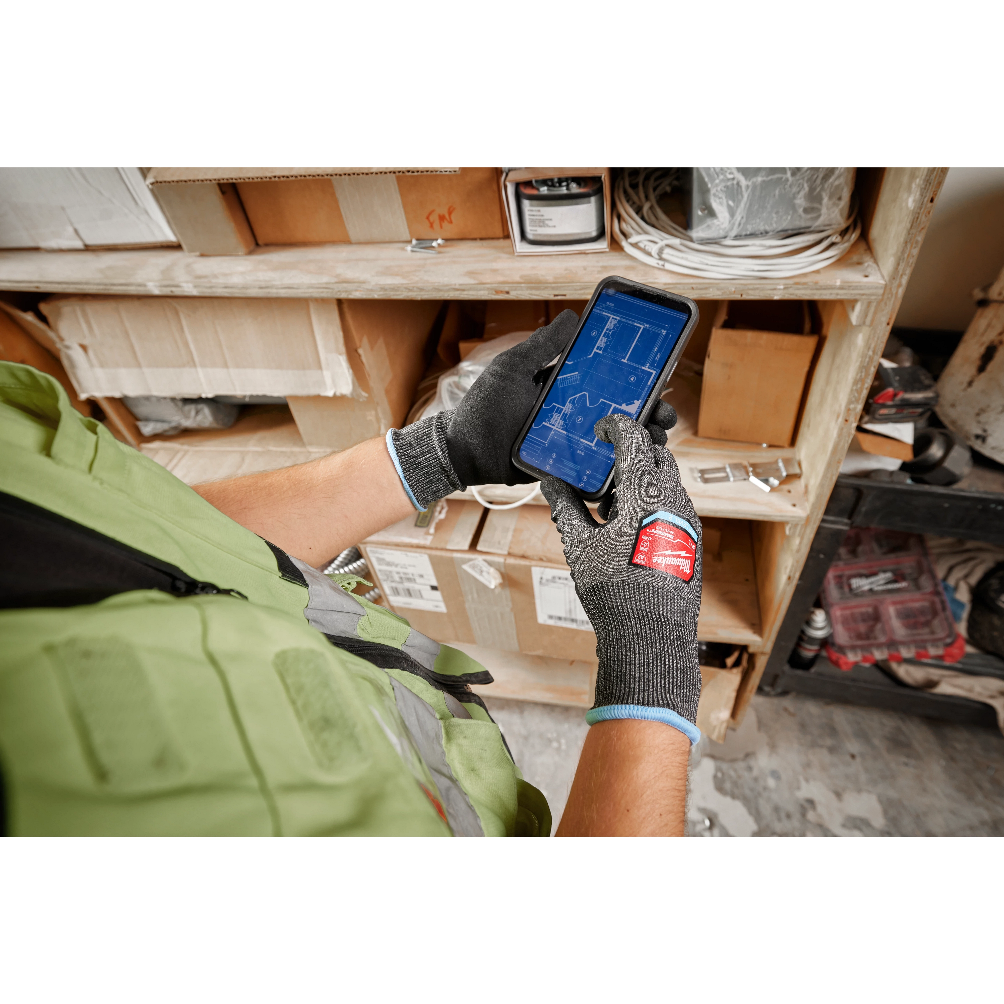 Person wearing Cut Level 2 High-Dexterity Nitrile Dipped Gloves - L, using a smartphone in a workshop with shelves and boxes.