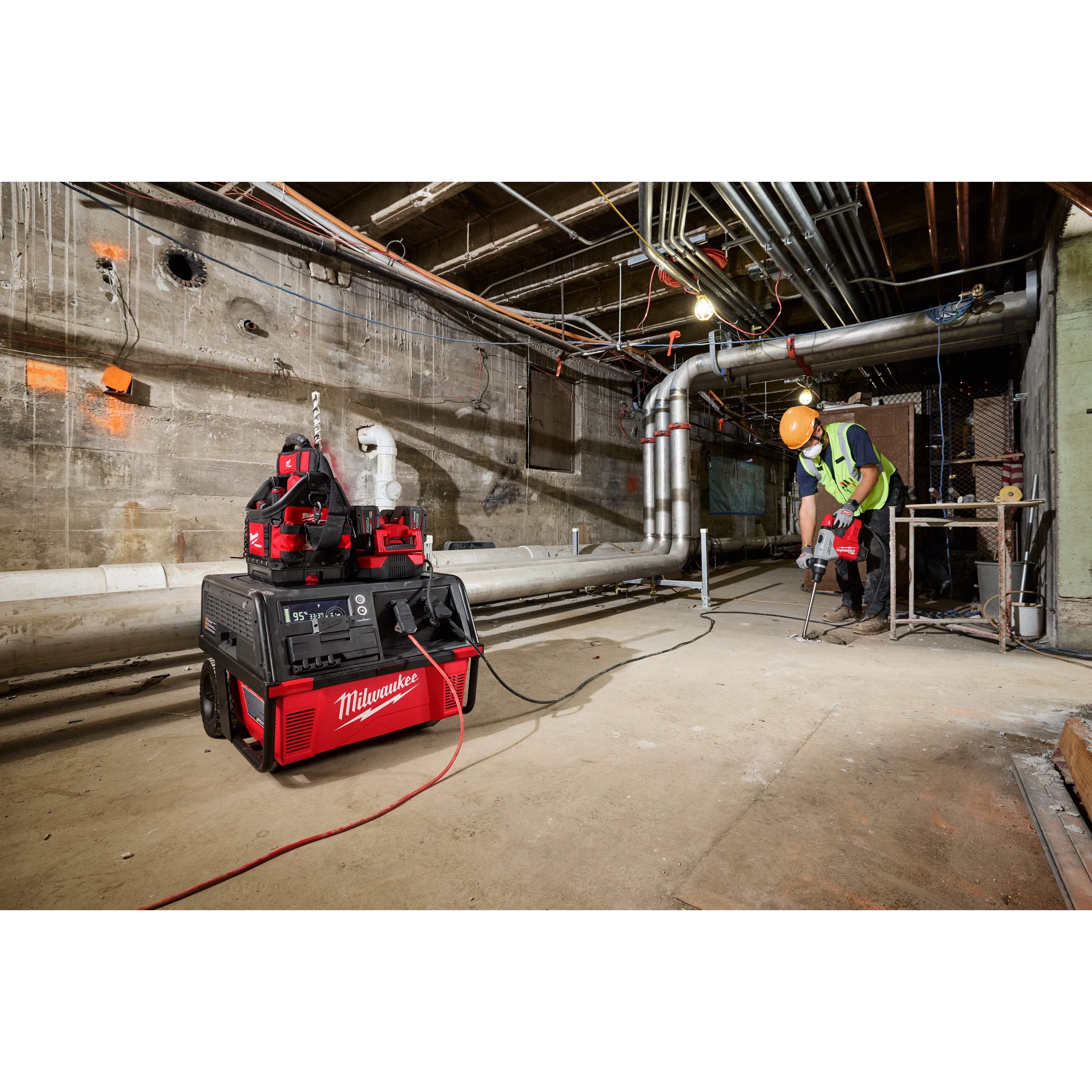 A worker using tools powered by the ROLL-ON™ 7200W/3600W 6.0kWh Power Supply in an industrial setting with exposed pipes and a concrete floor. The power supply is red and black, positioned near the worker and connected to their equipment.