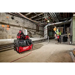 A worker using tools powered by the ROLL-ON™ 7200W/3600W 6.0kWh Power Supply in an industrial setting with exposed pipes and a concrete floor. The power supply is red and black, positioned near the worker and connected to their equipment.