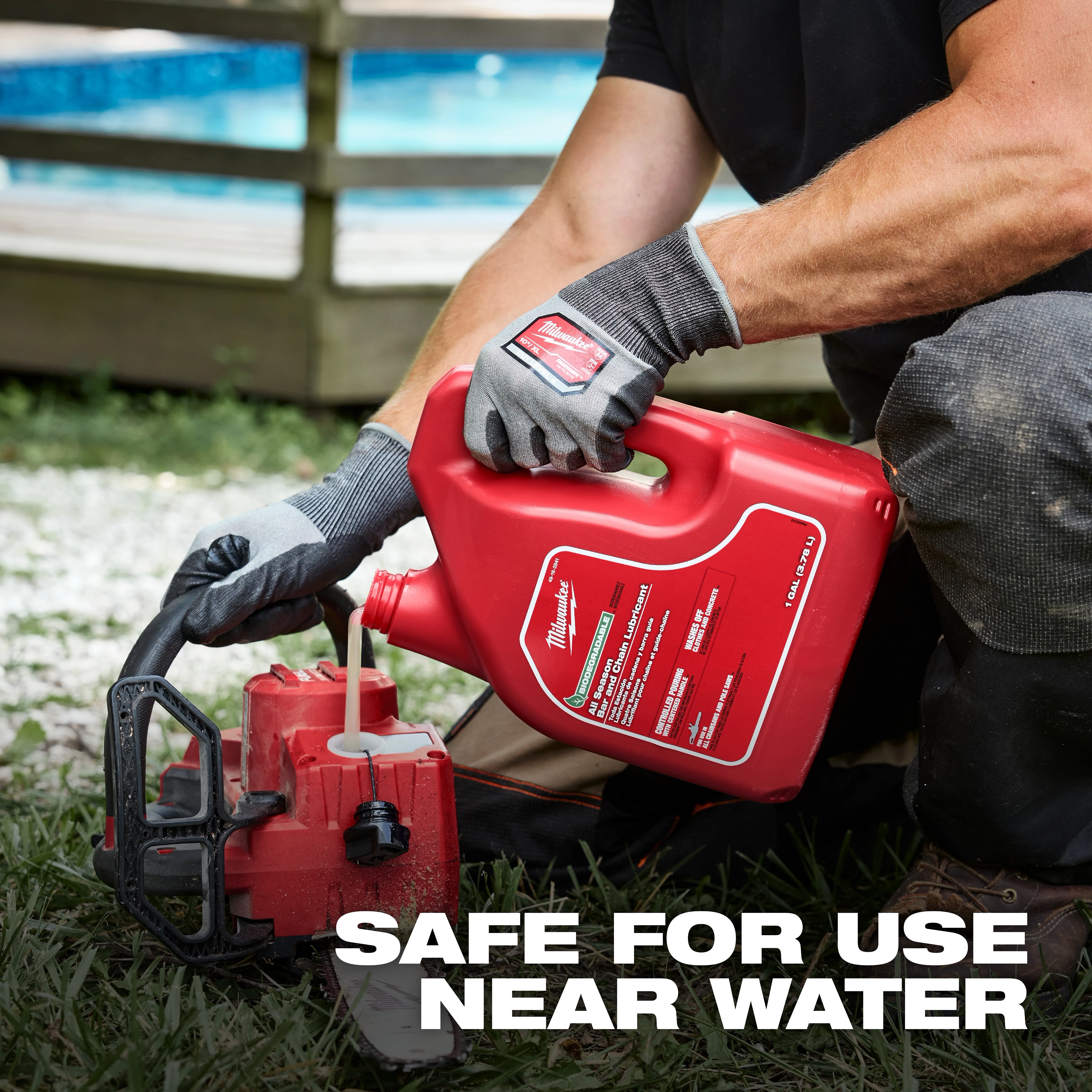 A person in work gloves and pants is pouring Biodegradable All Season Bar and Chain Lubricant from a large red container into a chainsaw's oil reservoir. The text on the image reads, "Safe for use near water." A blurred background includes a pool and wooden deck.