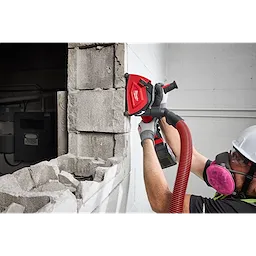 A worker uses a Milwaukee 5" Cutting Dust Shroud attached to a power tool to cut a concrete wall. The shroud is visible, connected to a hose for dust extraction.
