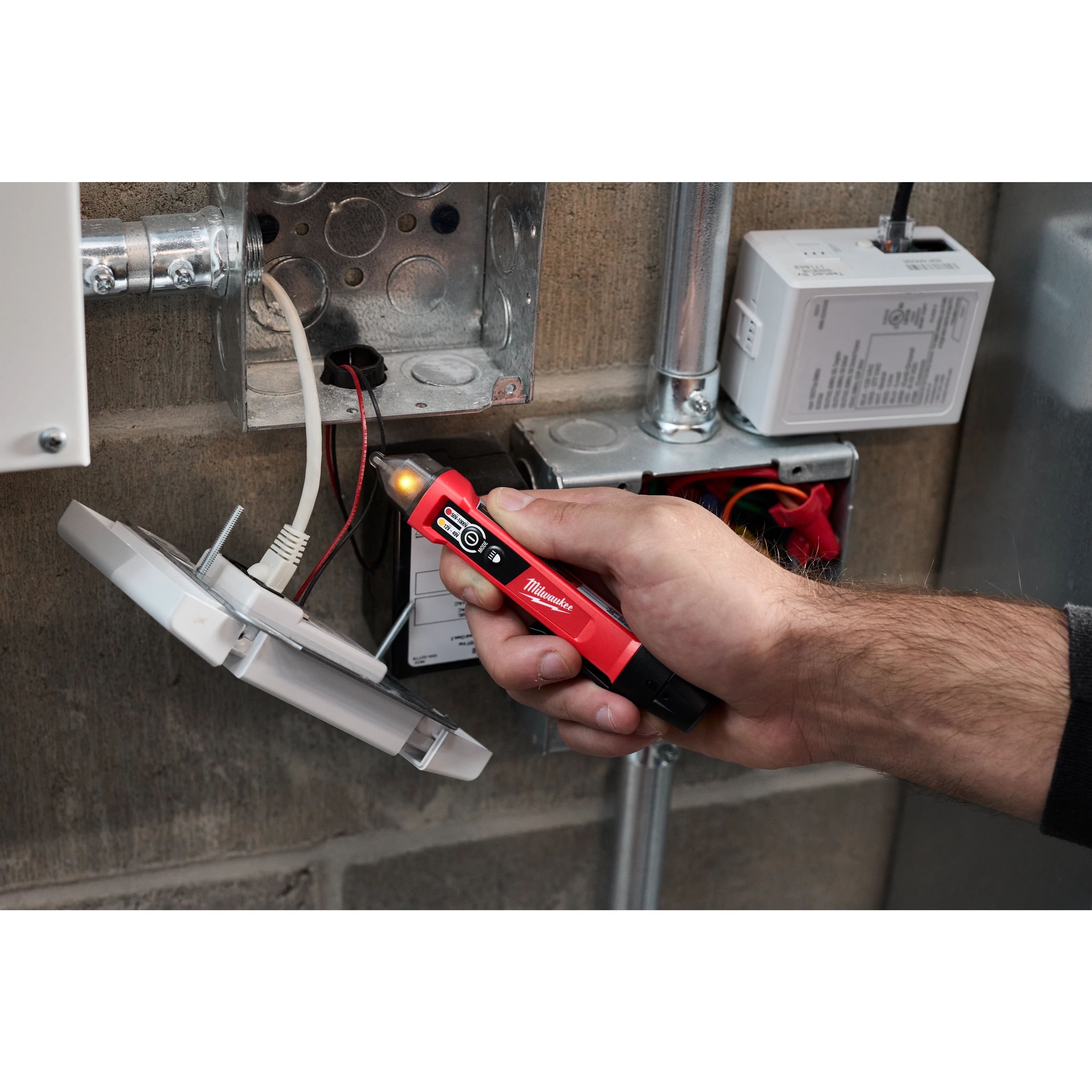 A person is using a Dual Range Voltage Detector w/ LED to check electrical connections in an unfinished metal junction box. The detector has a red and black design with an LED lighted tip indicating voltage presence.