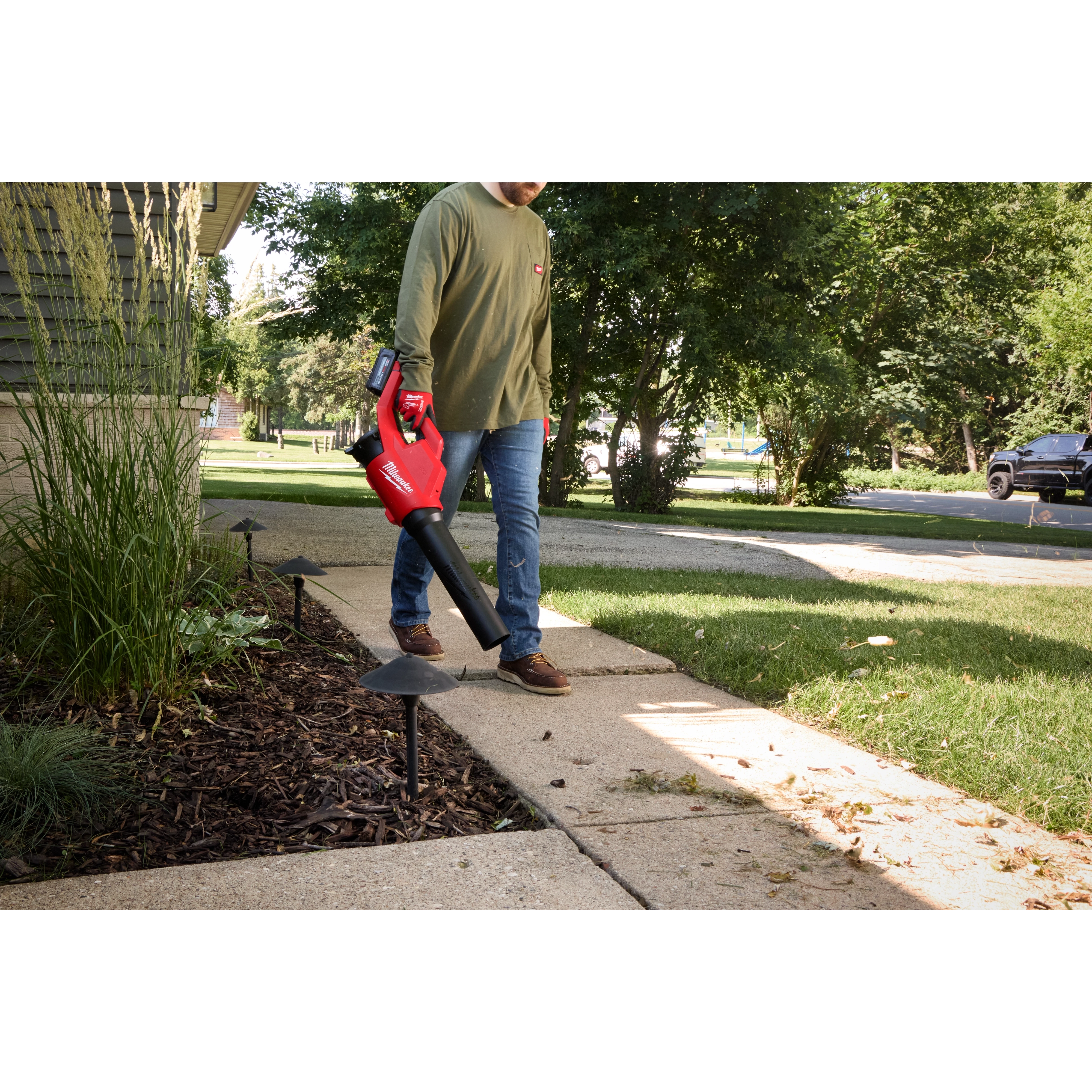 A person uses the M18™ Brushless Blower to clear leaves from a sidewalk adjacent to a landscaped garden. The blower is red and black with a comfortable grip handle. The scene takes place during the day in a suburban outdoor setting.