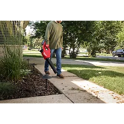 A person uses the M18™ Brushless Blower to clear leaves from a sidewalk adjacent to a landscaped garden. The blower is red and black with a comfortable grip handle. The scene takes place during the day in a suburban outdoor setting.