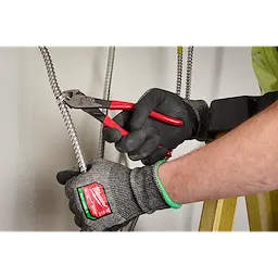 A person wearing gloves is using a pair of red-handled pliers to cut a metal conduit. The gloves are labeled "Milwaukee," and the individual is working near a wall with other cables visible. The person stands on a yellow ladder.