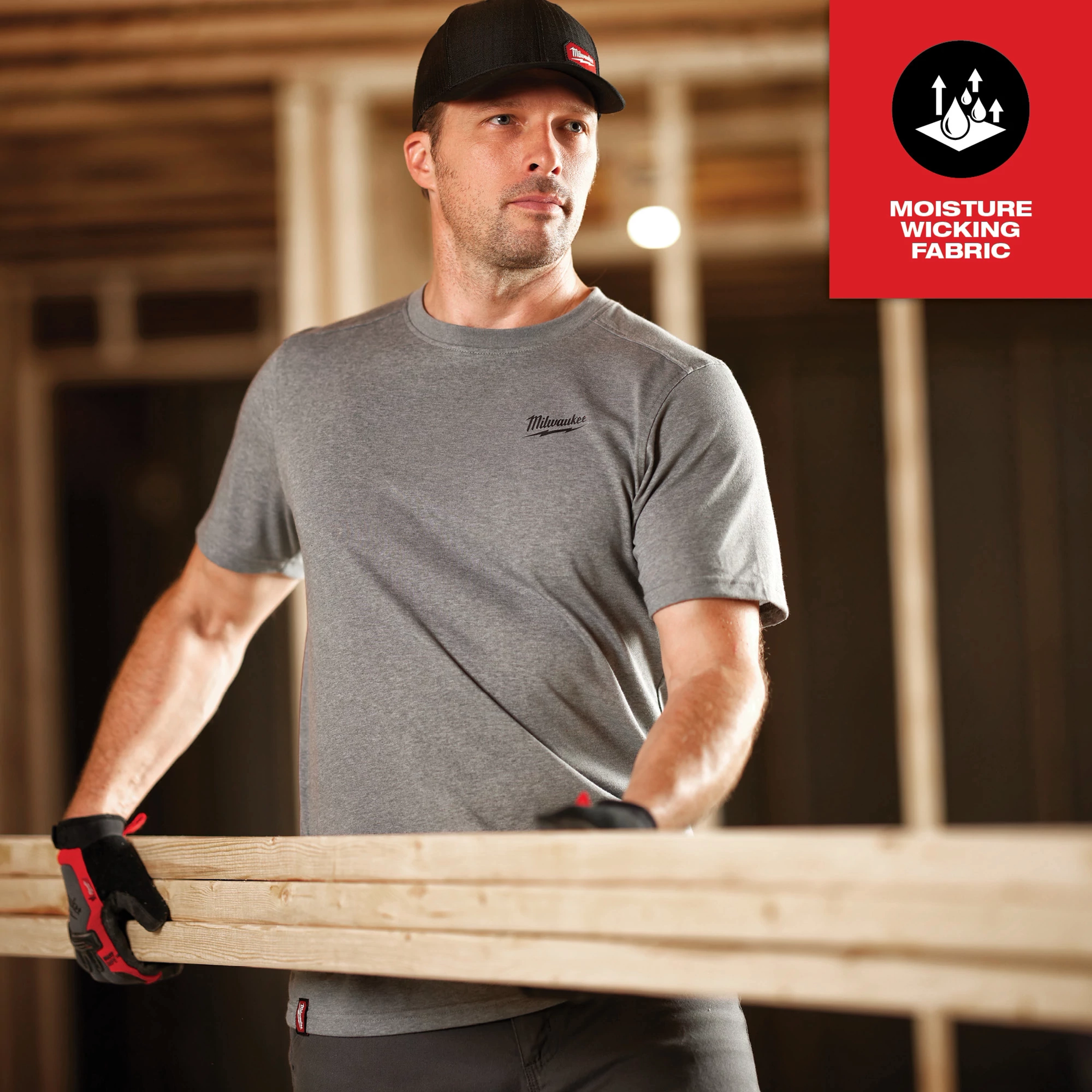 The image shows a man wearing the Hybrid Work Tee - Short Sleeve Feature Shot while holding wooden planks in a construction site.