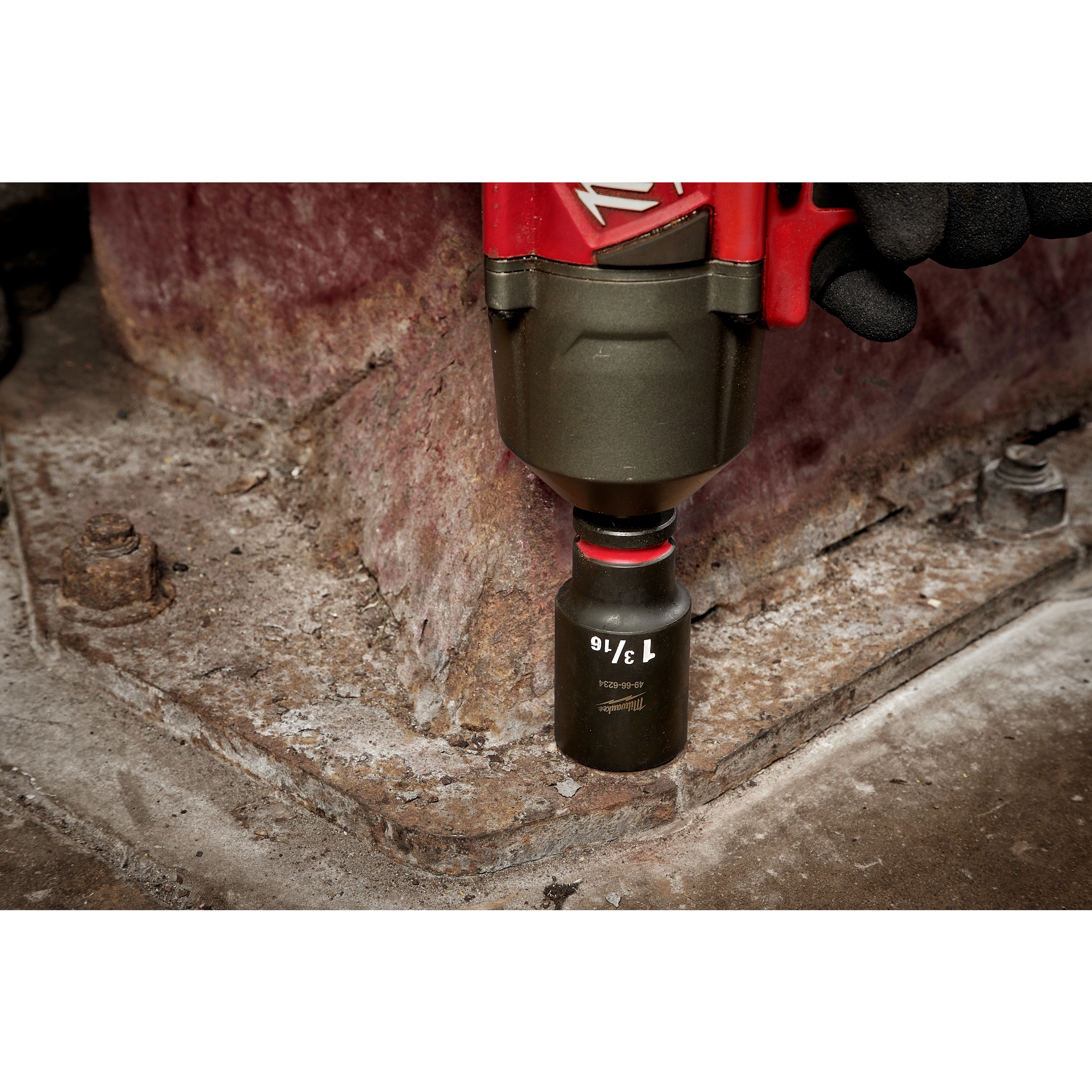 SHOCKWAVE Impact Duty™ 1/2" Drive 1-3/16" Deep 6 Point Socket, attached to a power tool, is being used by a gloved hand to work on a metal structure. The socket is black with white text indicating size, and the surrounding area shows a worn industrial surface.