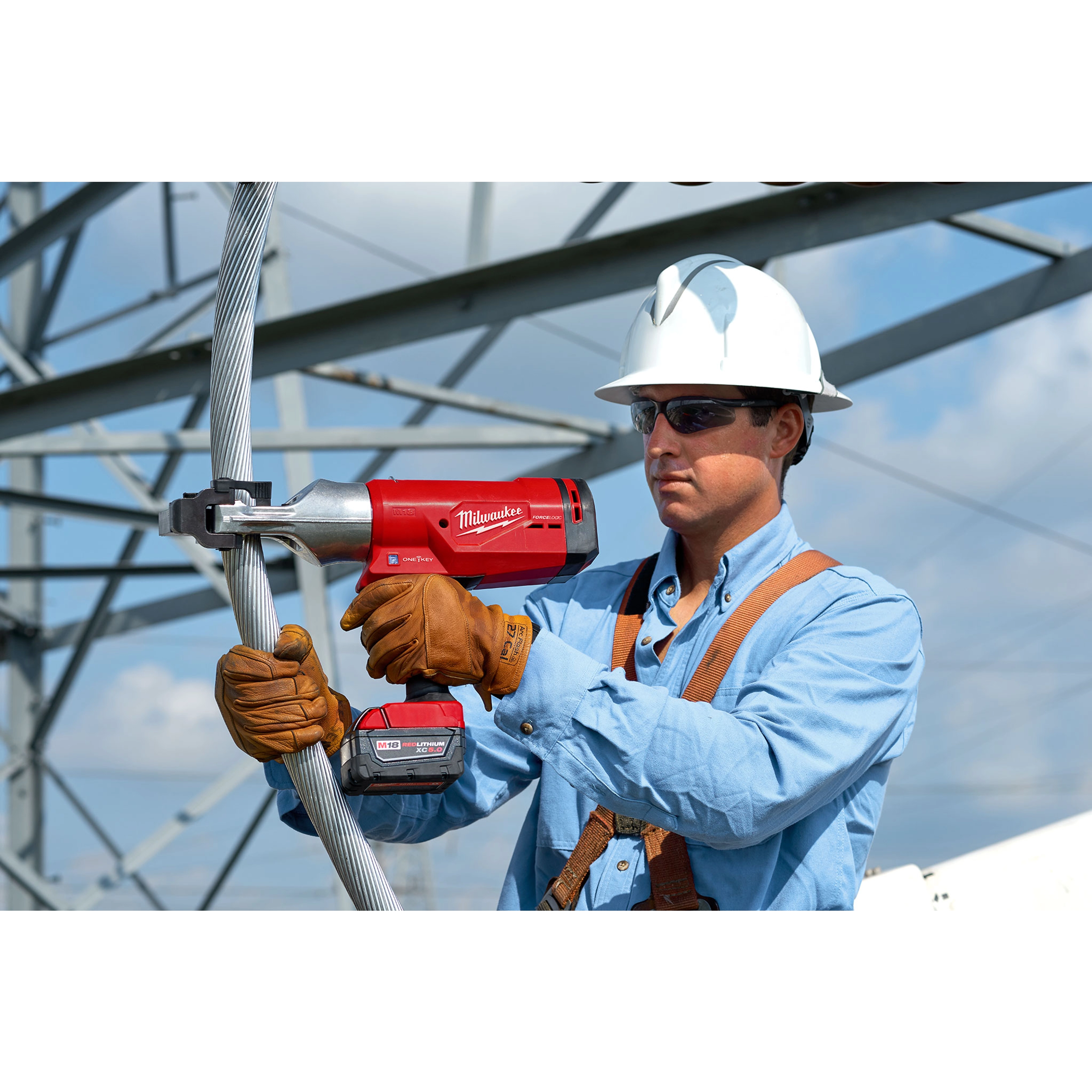 A worker uses the FORCELOGIC™ M18™ 1590 ACSR Cutter Kit to cut a thick metal cable. He is wearing a hard hat, safety gloves, and blue work clothing with a harness.