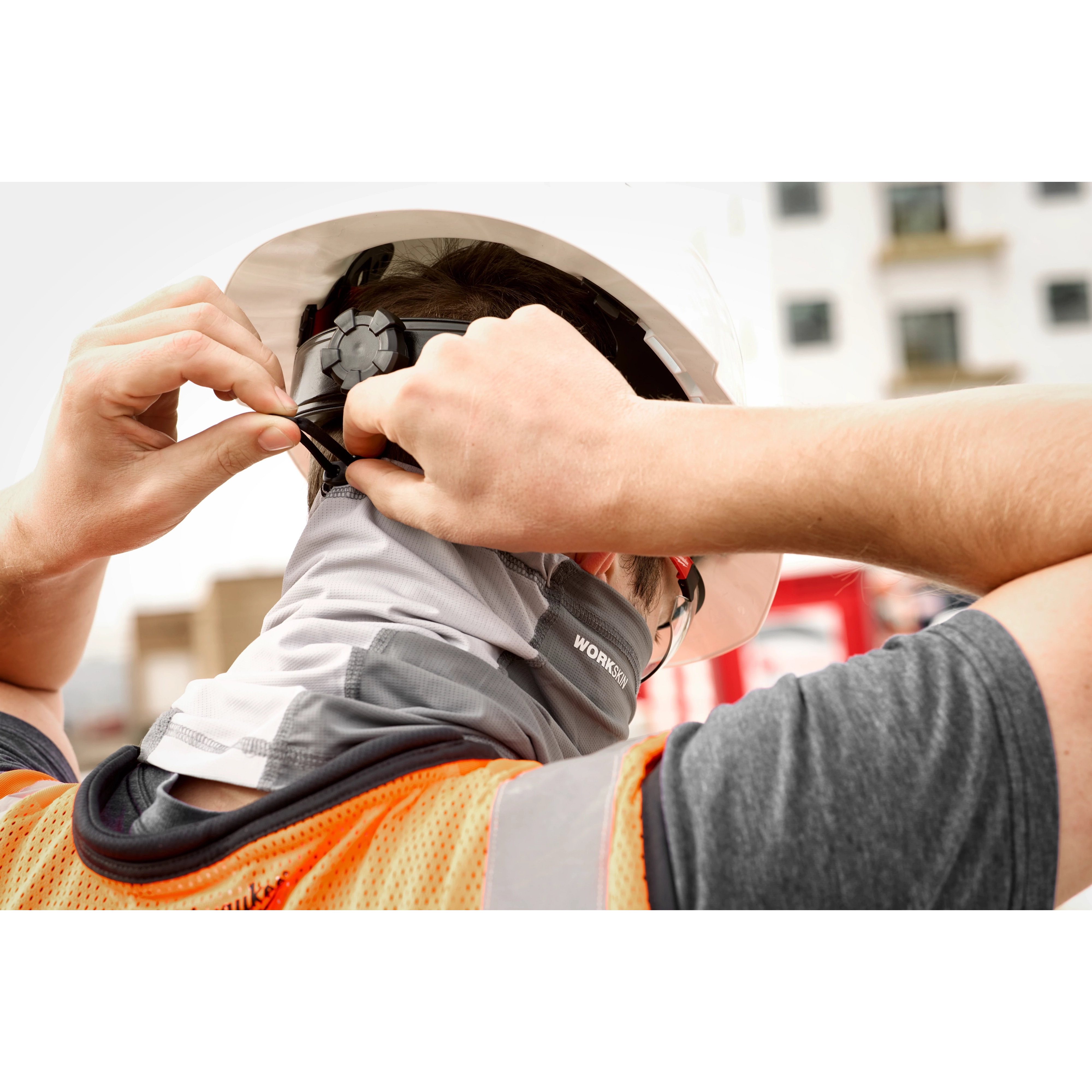 Person wearing a hard hat adjusting the WORKSKIN Performance Neck Gaiter at a construction site, also wearing an orange safety vest.