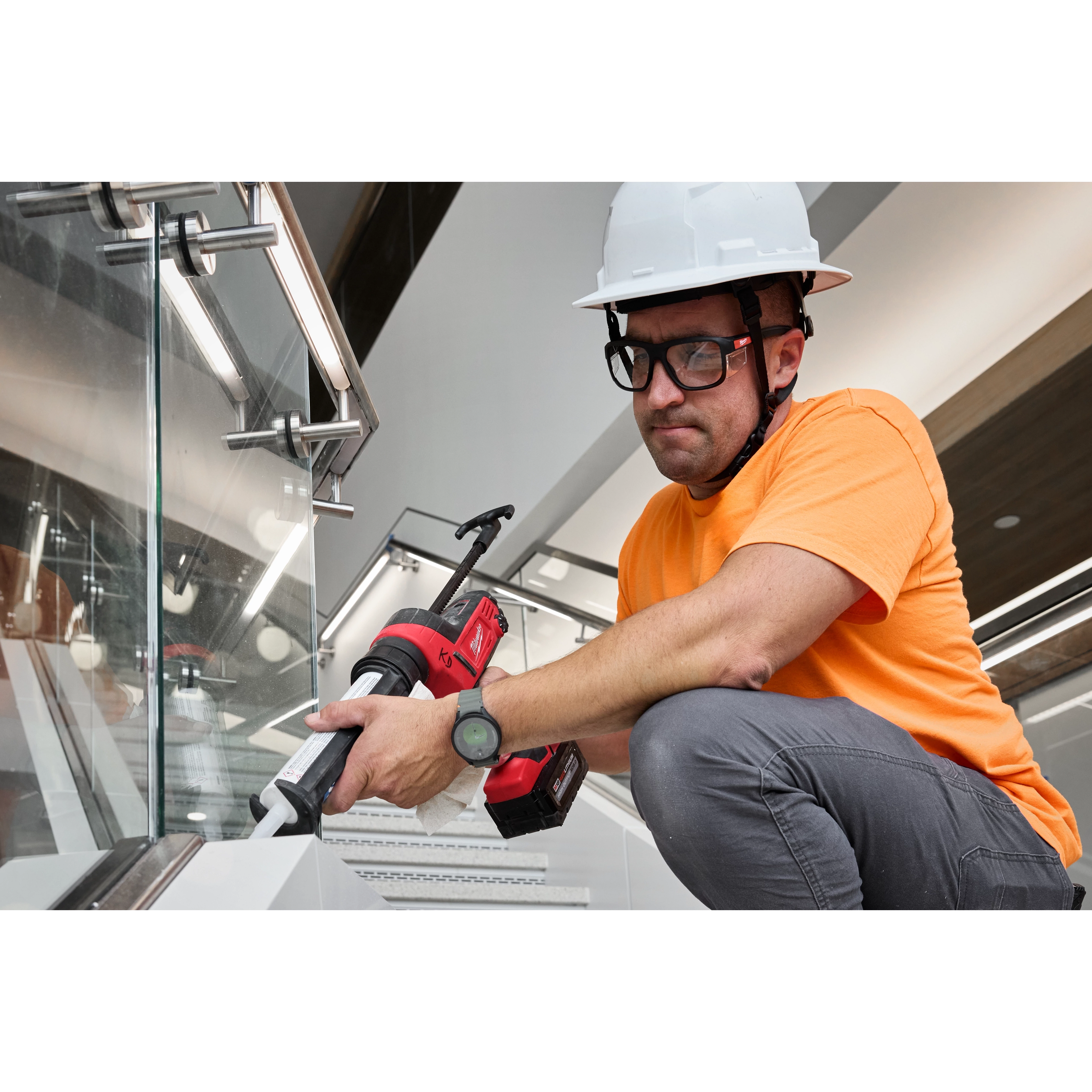 A worker in an orange shirt and white hard hat uses a power caulking gun on a stairwell. The worker is wearing Full Frame Safety Glasses with Removable Side Shields – Clear Anti-Scratch Lenses for eye protection. The surroundings include metal railings and glass panels.