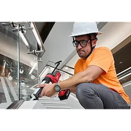 A worker in an orange shirt and white hard hat uses a power caulking gun on a stairwell. The worker is wearing Full Frame Safety Glasses with Removable Side Shields – Clear Anti-Scratch Lenses for eye protection. The surroundings include metal railings and glass panels.