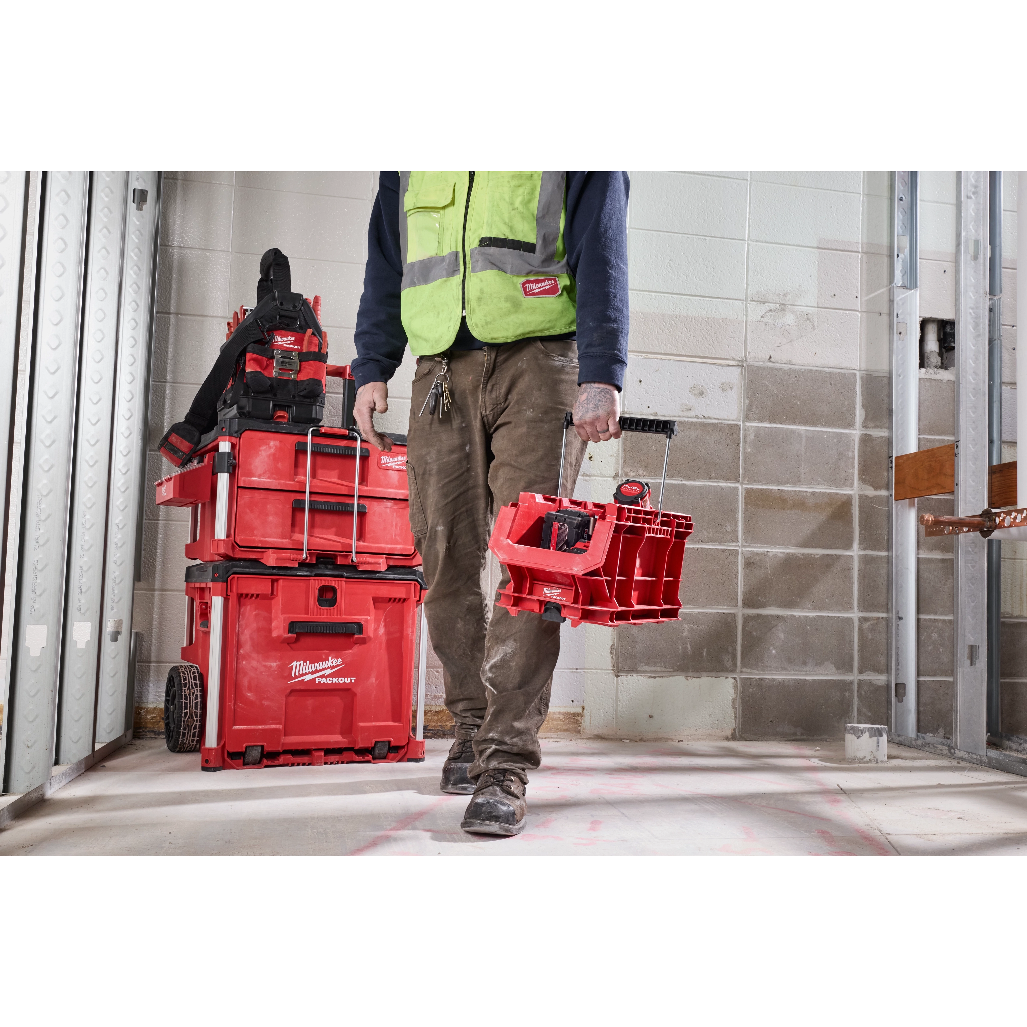 A person carrying a PACKOUT Compact Crate in a construction setting, with a stacked set of red PACKOUT toolboxes in the background.