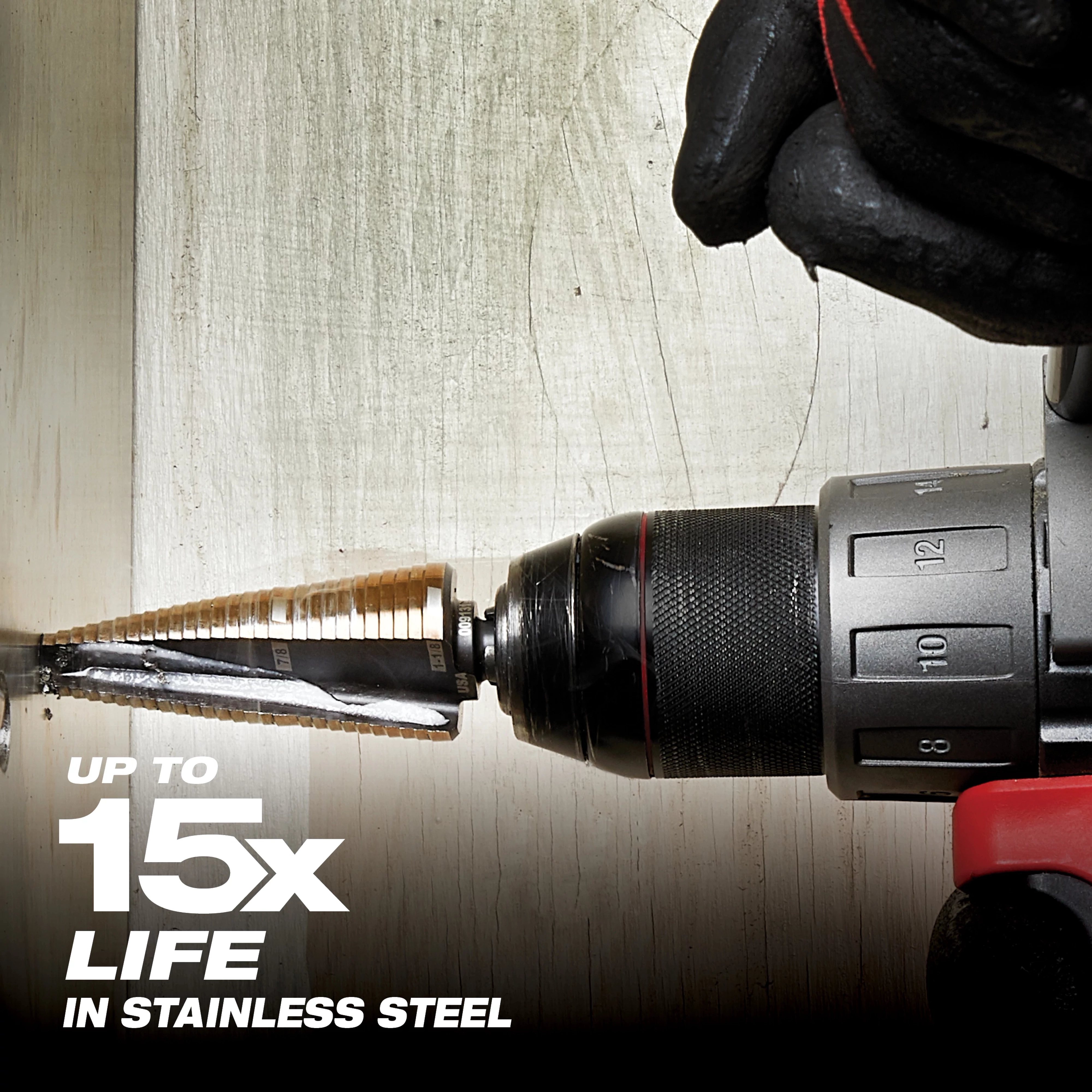 A gloved hand operates a power drill with a Cobalt%20Step%20Bit attached, drilling into stainless steel. The text "Up to 15x life in stainless steel" appears in the lower-left corner, indicating the product's durability.