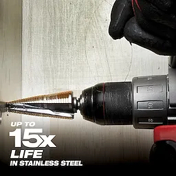 A gloved hand operates a power drill with a Cobalt%20Step%20Bit attached, drilling into stainless steel. The text "Up to 15x life in stainless steel" appears in the lower-left corner, indicating the product's durability.