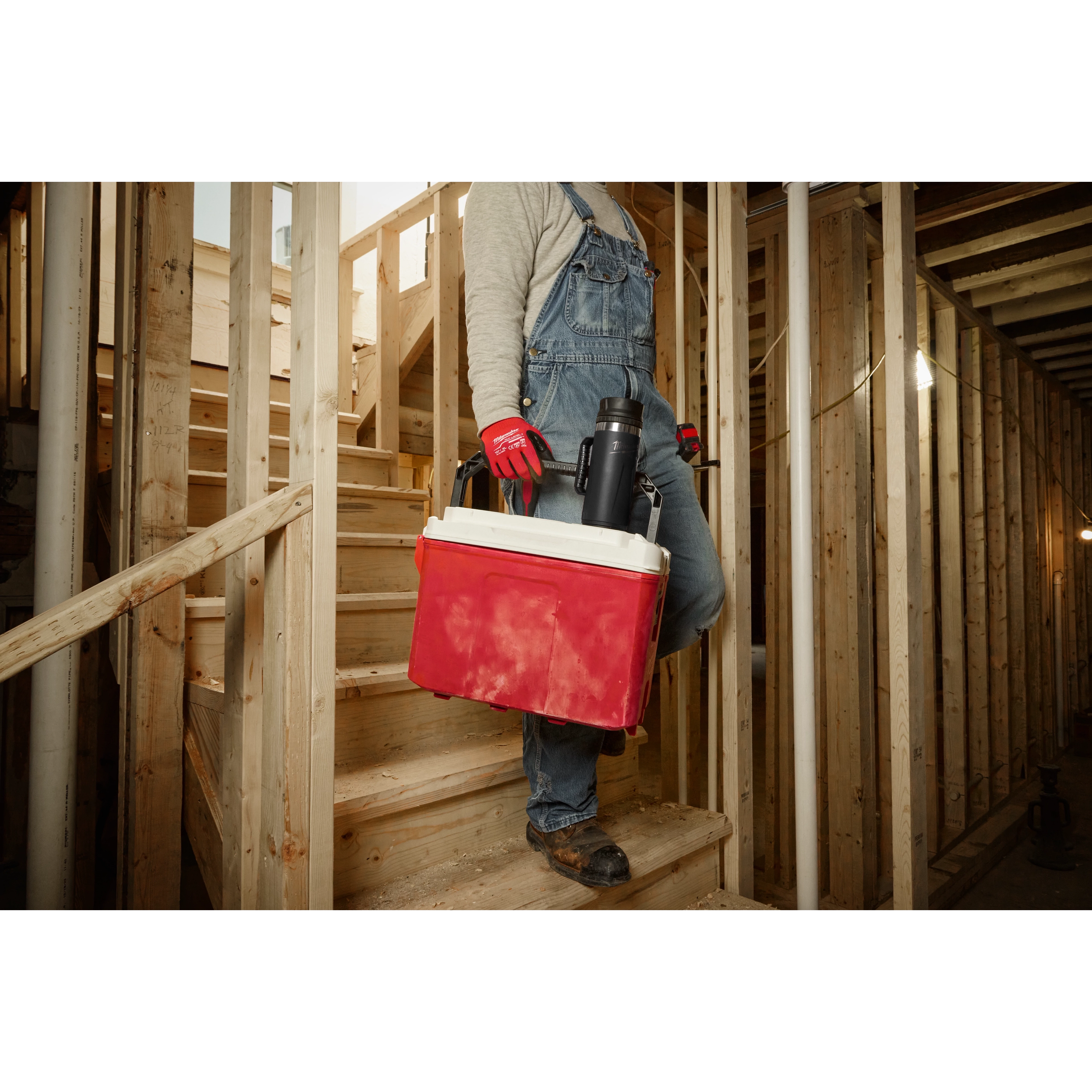 Worker in overalls carrying a red cooler and a PACKOUT 18oz Insulated Mug with Sip Lid, descending wooden construction stairs.