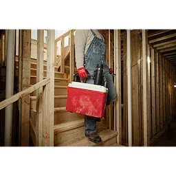 Worker in overalls carrying a red cooler and a PACKOUT 18oz Insulated Mug with Sip Lid, descending wooden construction stairs.