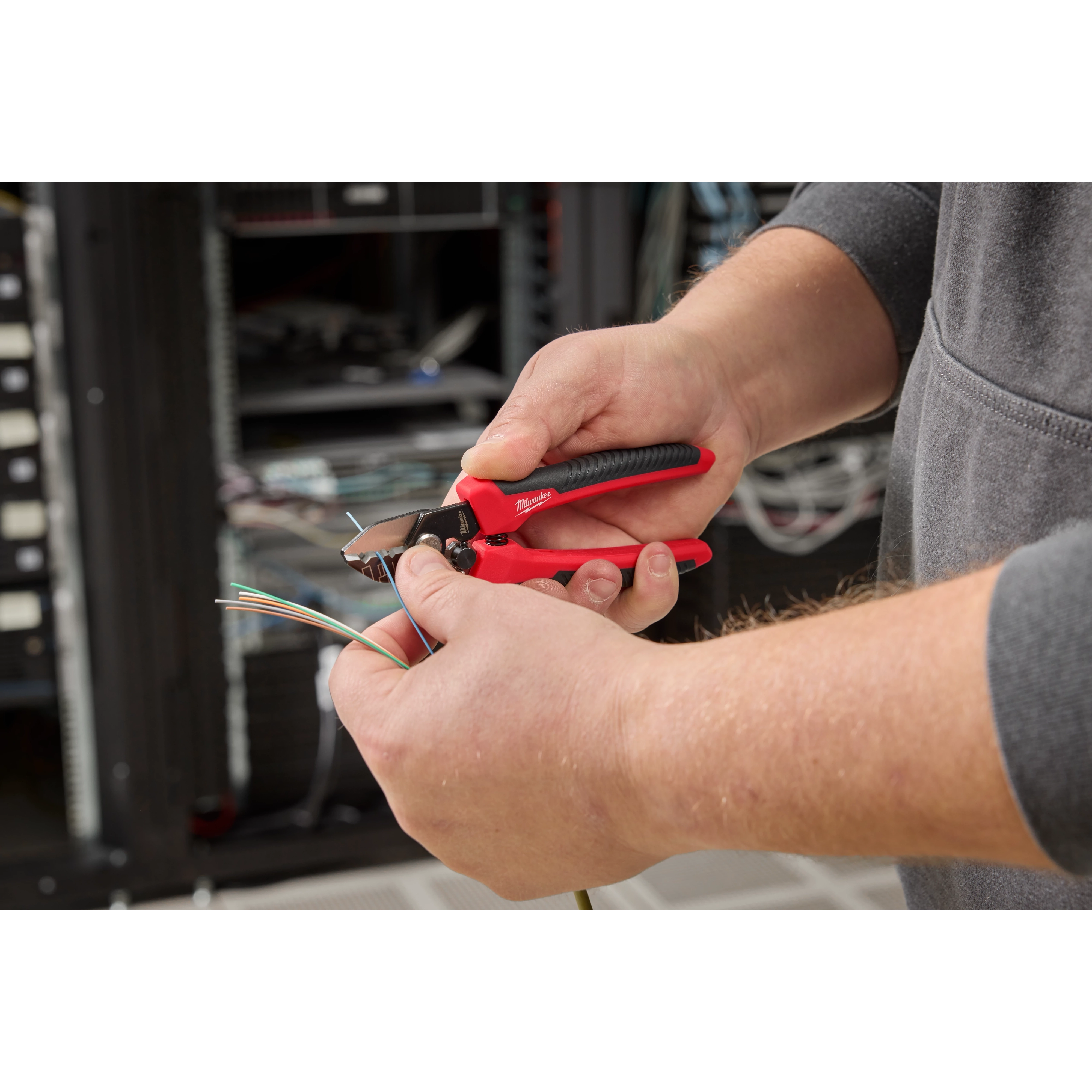 A person uses a red and black Tri-Hole Fiber Stripper to strip the insulation from multiple colored fiber optic cables. The background shows networking equipment and cables.