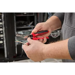 A person uses a red and black Tri-Hole Fiber Stripper to strip the insulation from multiple colored fiber optic cables. The background shows networking equipment and cables.