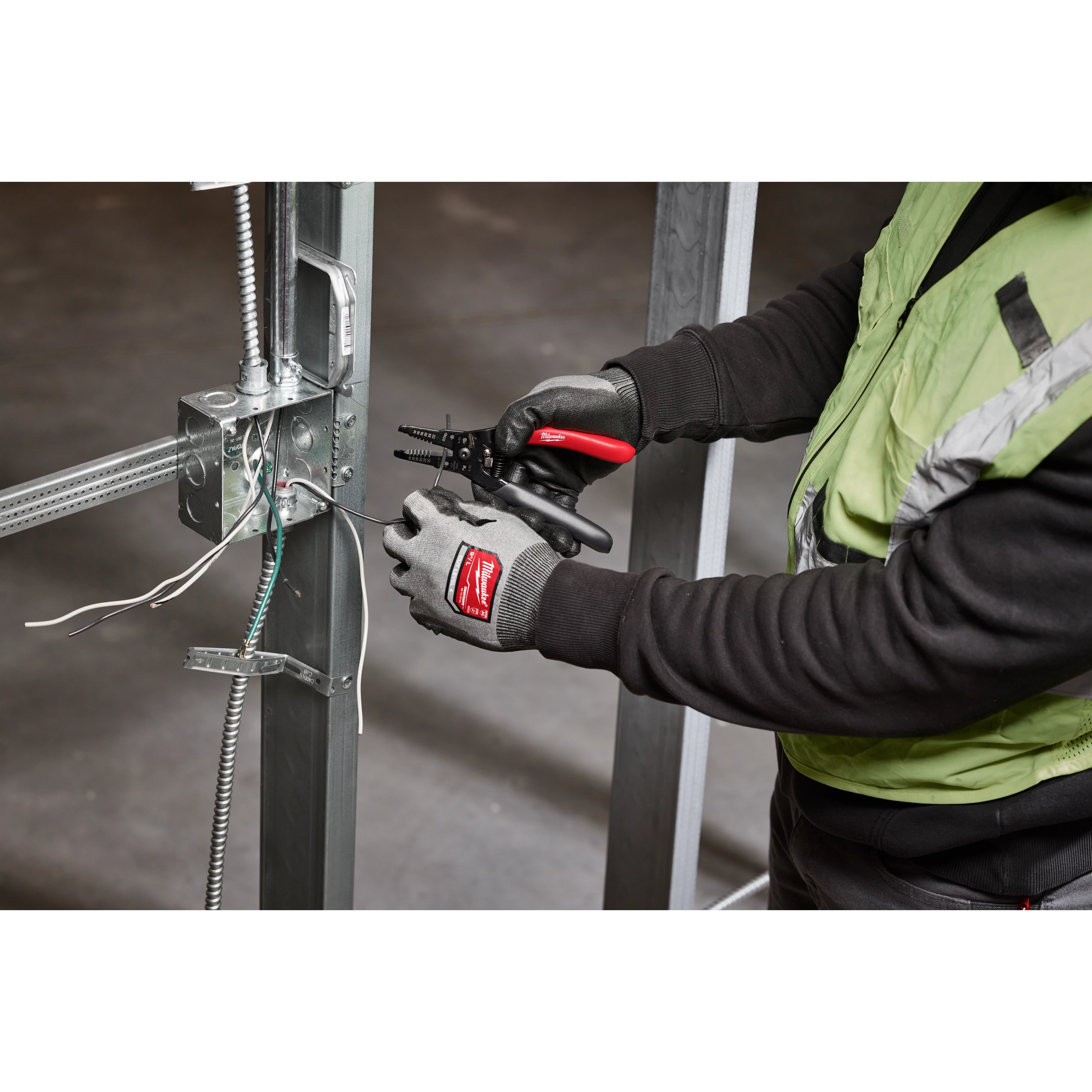 A person wearing gloves uses a 20-32% AWG Low Voltage Dipped Grip Wire Stripper & Cutter to strip wires in an industrial setting with metal framework.