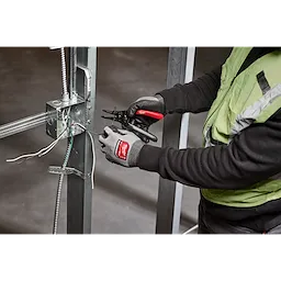 A person wearing gloves uses a 20-32% AWG Low Voltage Dipped Grip Wire Stripper & Cutter to strip wires in an industrial setting with metal framework.