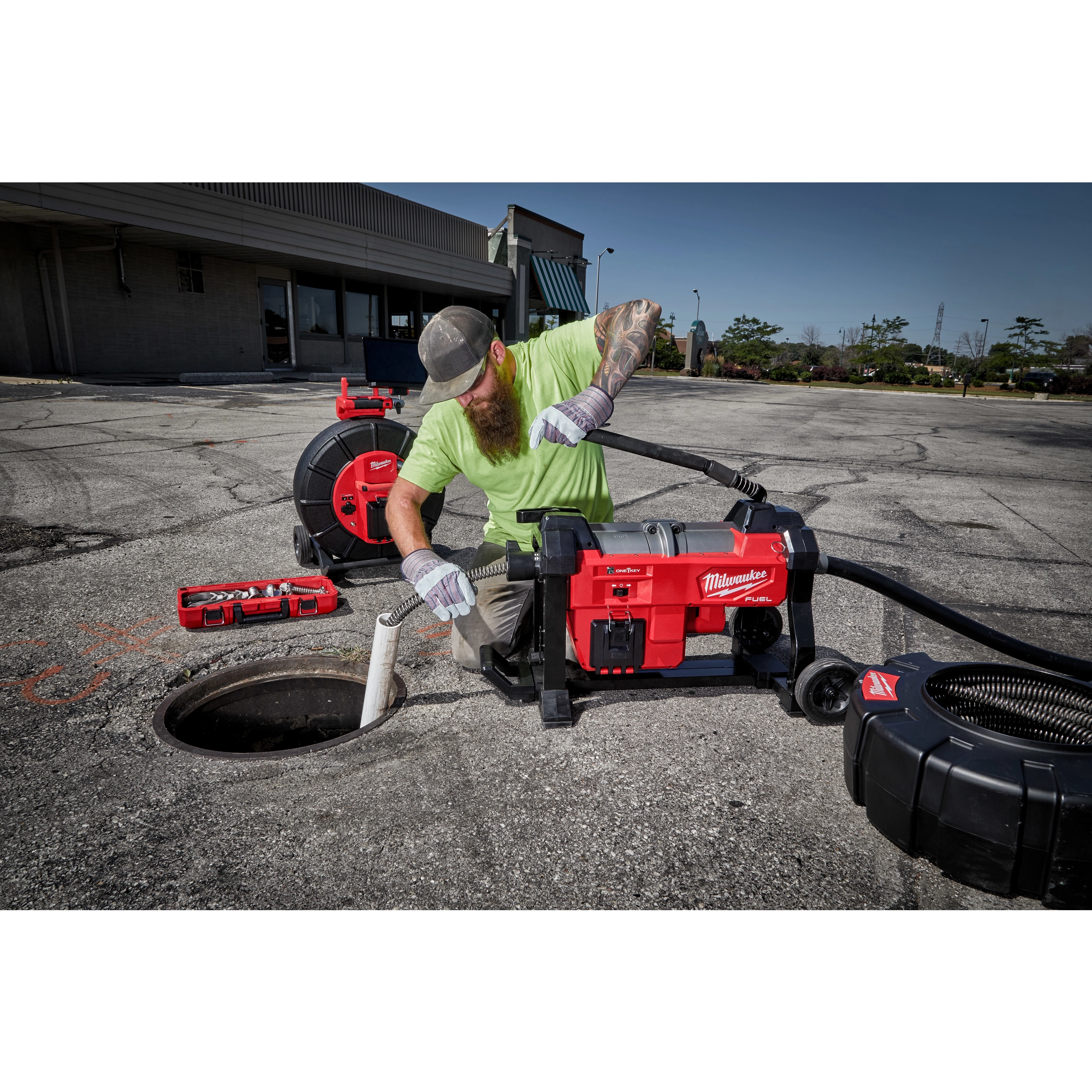A worker in a green shirt and cap operates a 200’ Stiff Modular Pipeline Inspection Reel by Milwaukee in an outdoor setting. The reel is being used to inspect an underground pipeline, with various tools and equipment visible nearby. The area is a paved surface near a building.